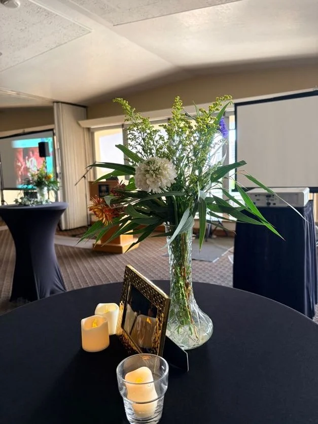 A table decorated with a glass vase of flowers, a framed photograph, and LED candles in a conference room or banquet hall.