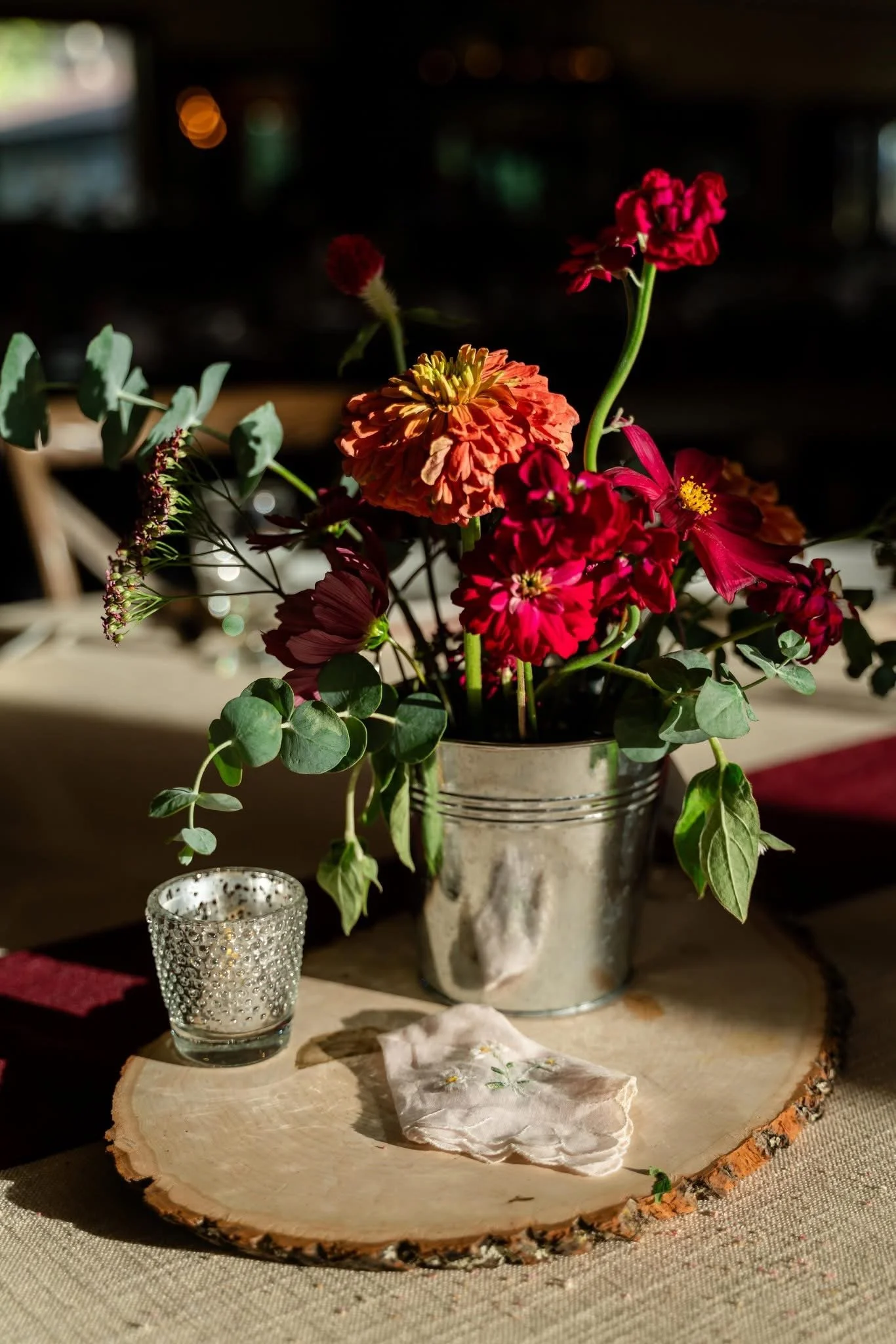 A floral arrangement featuring red and orange flowers in a metal bucket on a wooden slice, with a small decorative candle holder and a piece of cloth on a table.