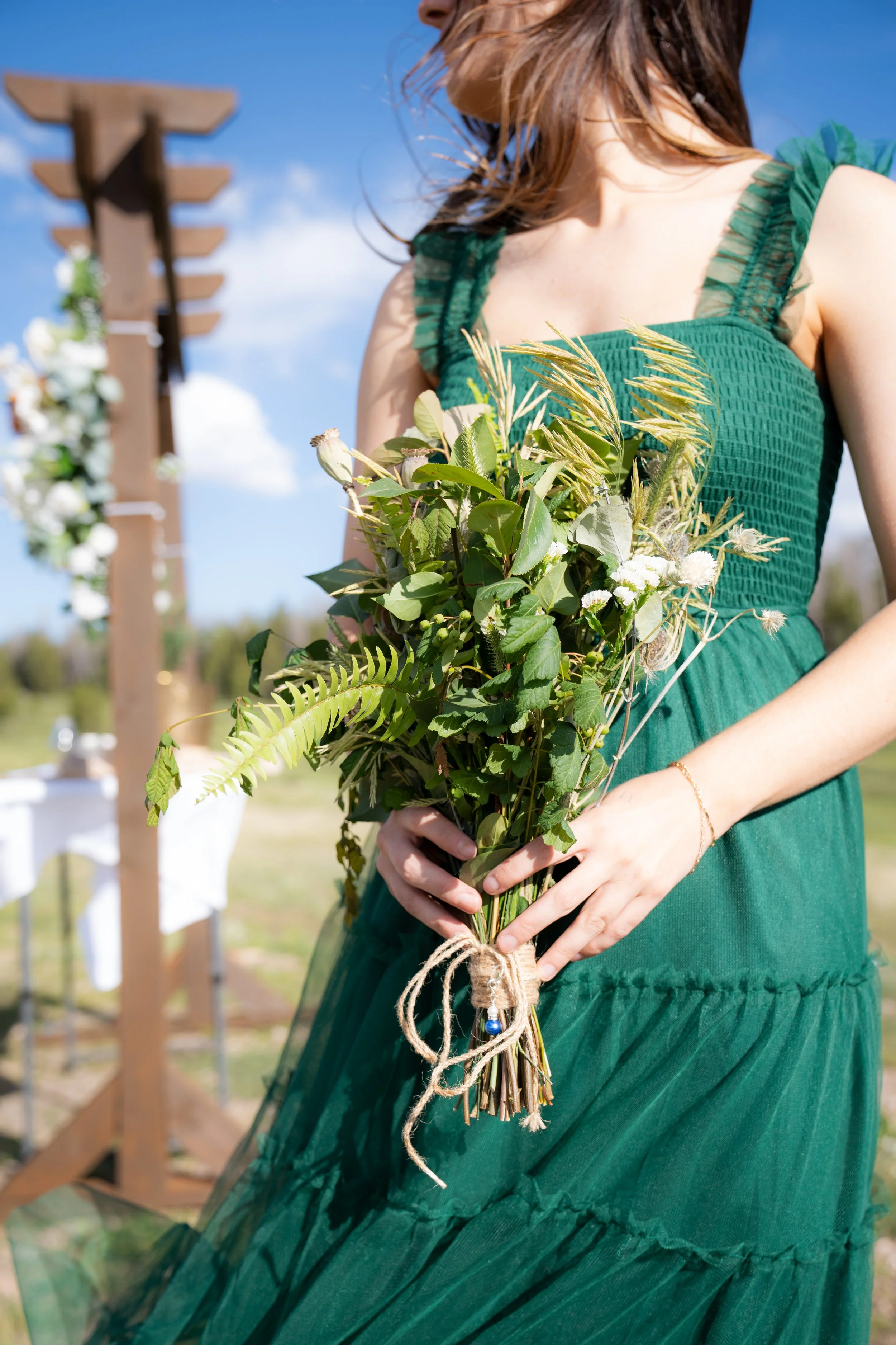A woman in a green dress holding a bouquet of various green and white flowers and foliage, outdoors on a sunny day with a blue sky and a wooden wedding arch in the background.