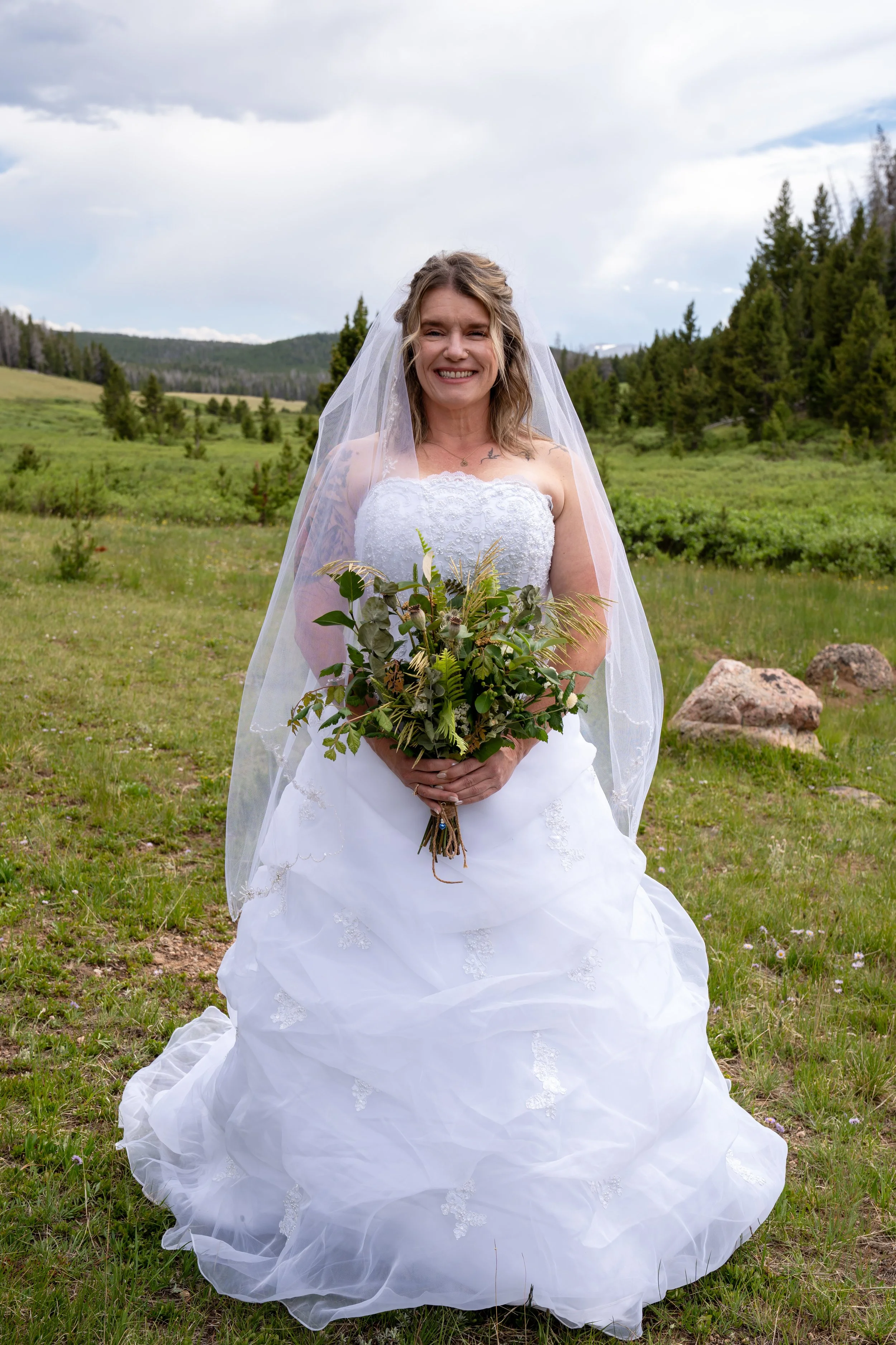 A smiling woman in a wedding dress with a veil, standing outdoors in a grassy field with mountains and trees in the background, holding a bouquet of green foliage.