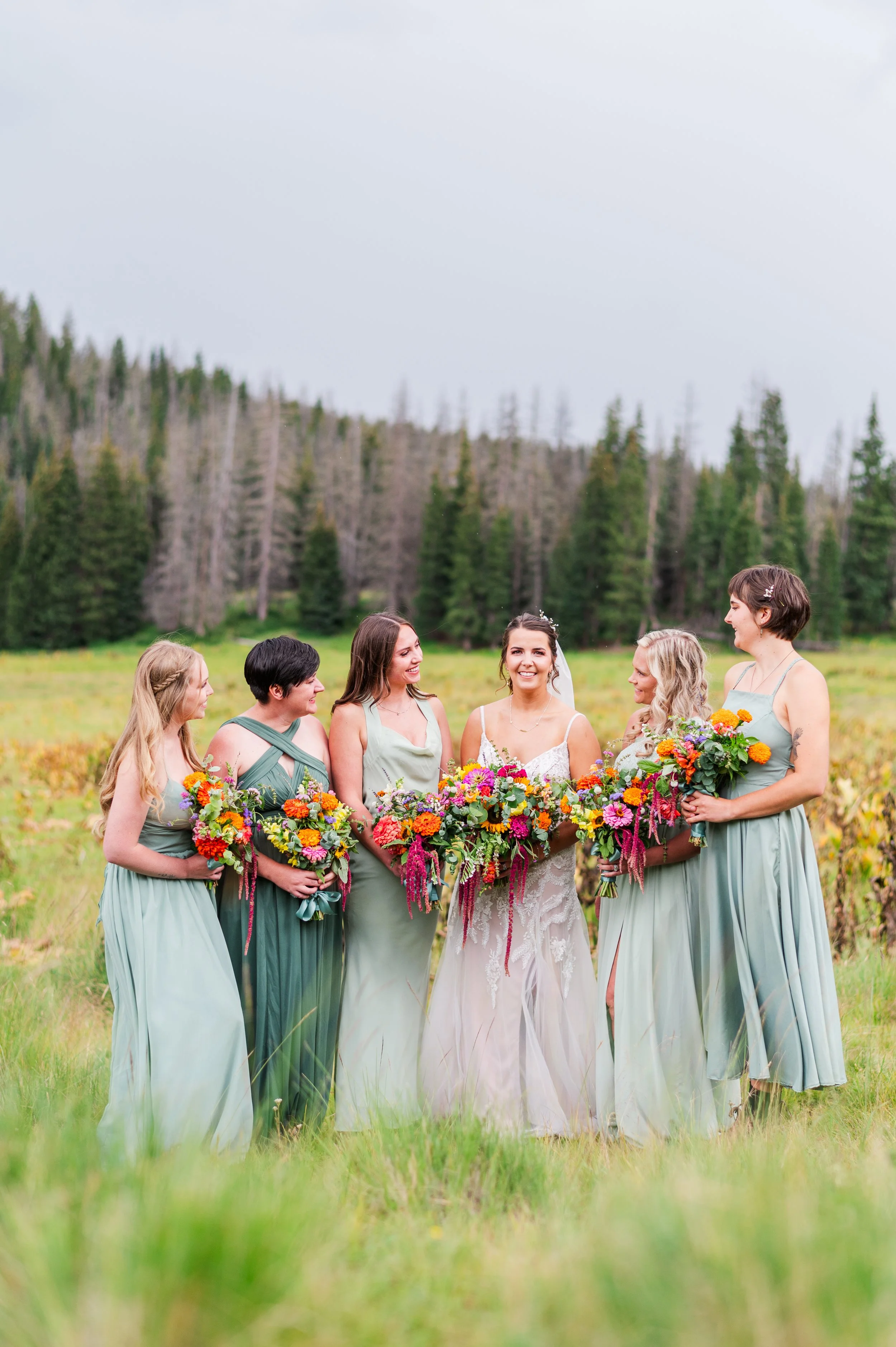 A group of women dressed in pastel-colored dresses, holding vibrant floral bouquets, standing together outdoors on a grassy field with a forested hillside in the background, celebrating a wedding.