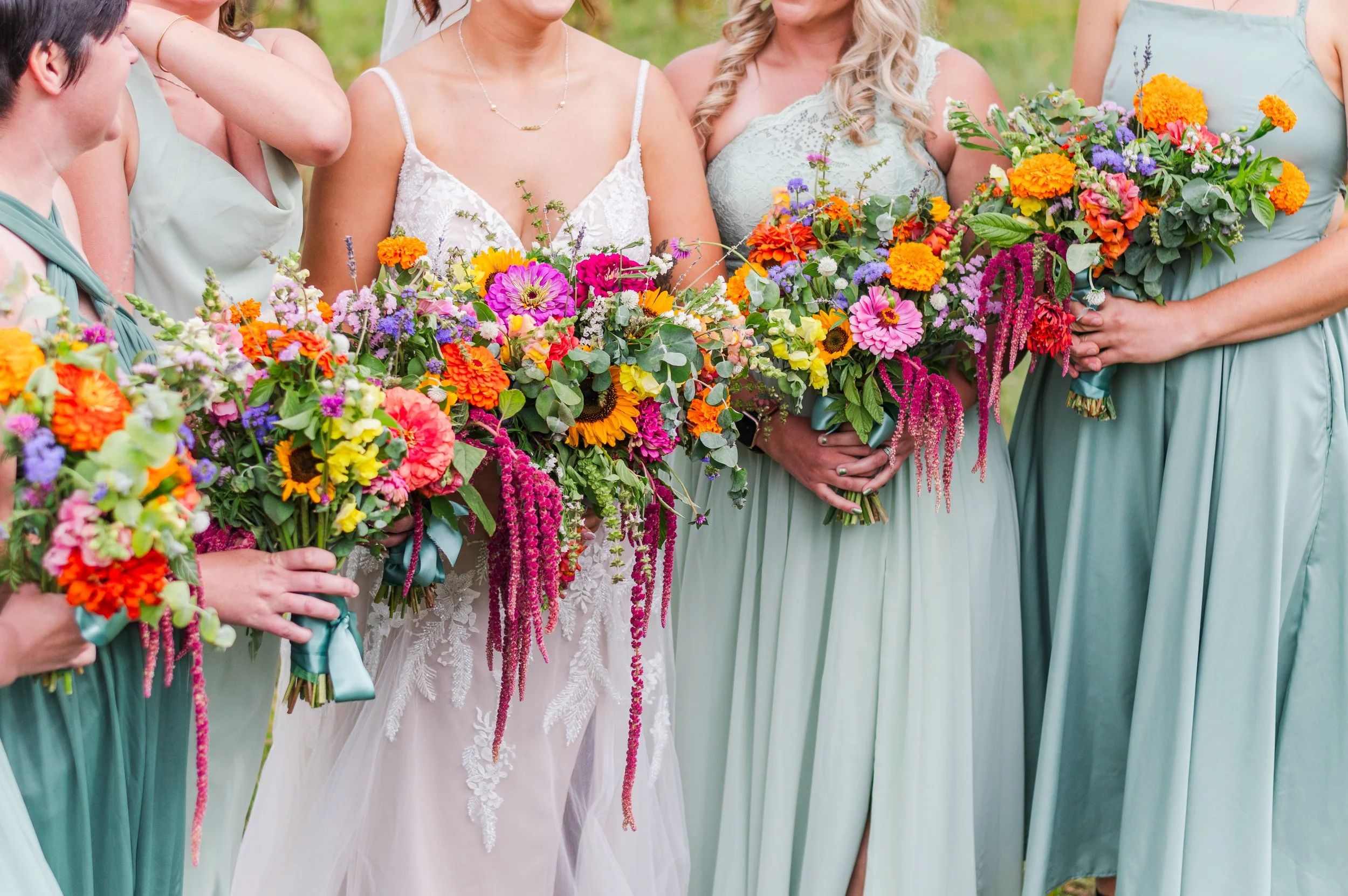 Women in mint green dresses and a bride in a white lace wedding gown holding colorful flower bouquets at an outdoor wedding.
