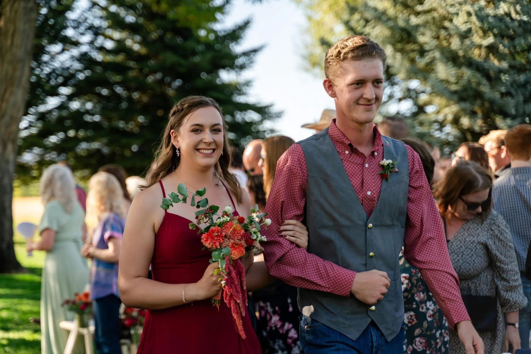 A young woman in a burgundy dress holding a colorful bouquet of flowers, walking arm-in-arm with a young man in a red checkered shirt and gray vest, amidst a crowd outdoors on a sunny day, possibly at a wedding or celebration.