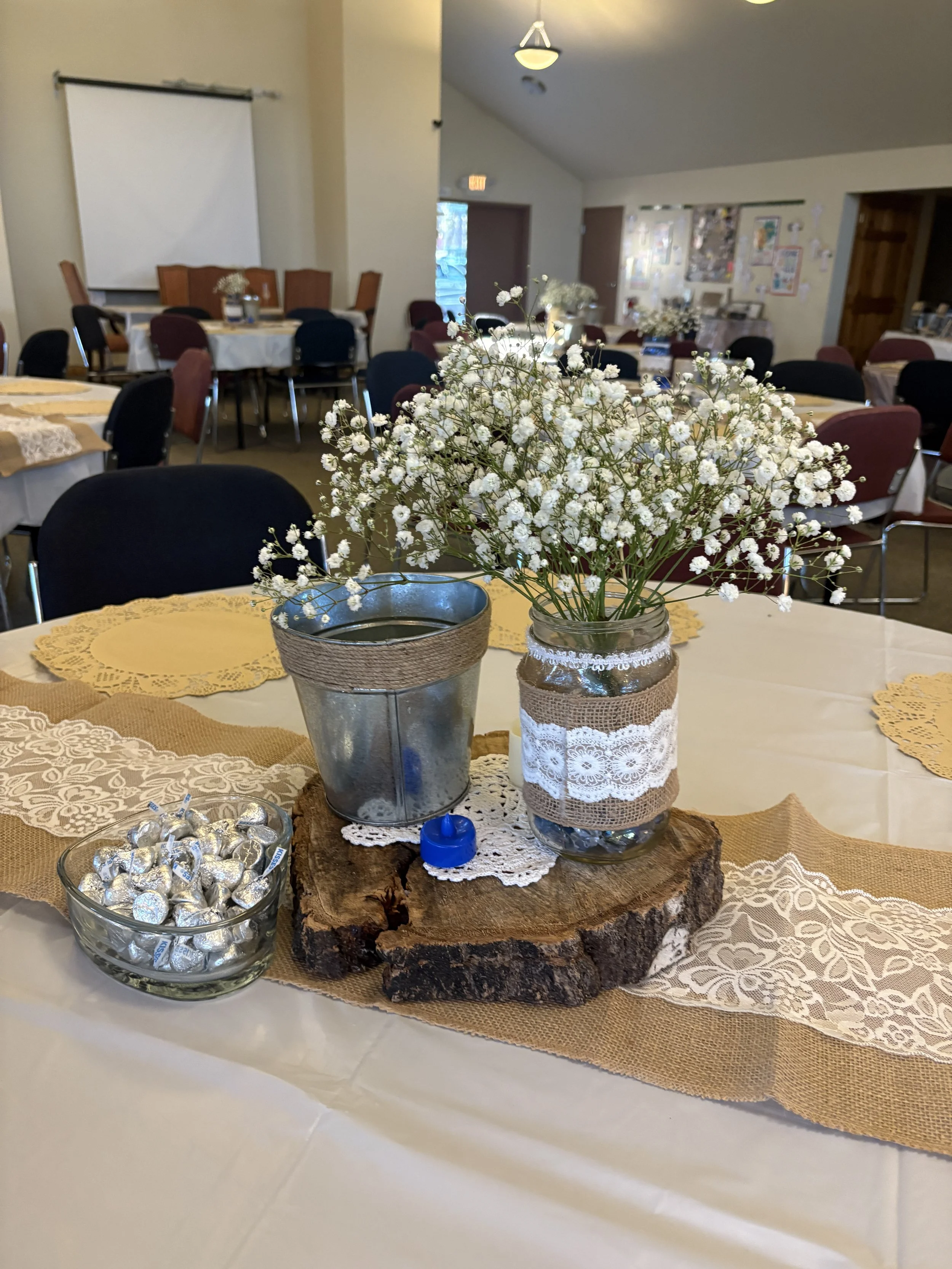 A centerpiece with white baby's breath flowers in a glass jar wrapped with lace and burlap, placed on a round wooden slice on a table with a burlap and lace table runner, alongside a bowl of small foil-wrapped chocolates and a small blue plastic toy.