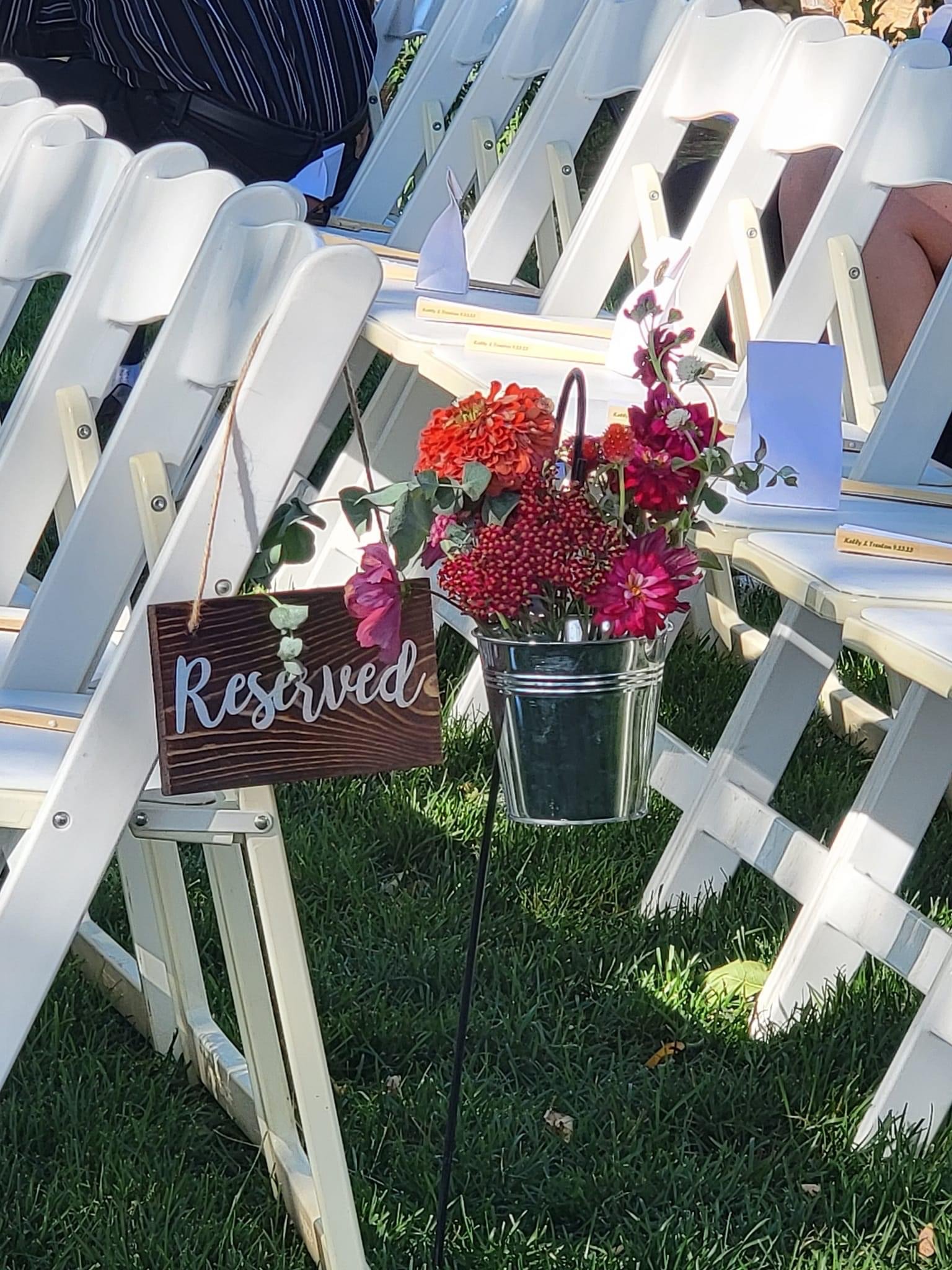 A row of white outdoor chairs with reserved signs, flowers in a vase, and a bouquet of plants, set on a grassy area.