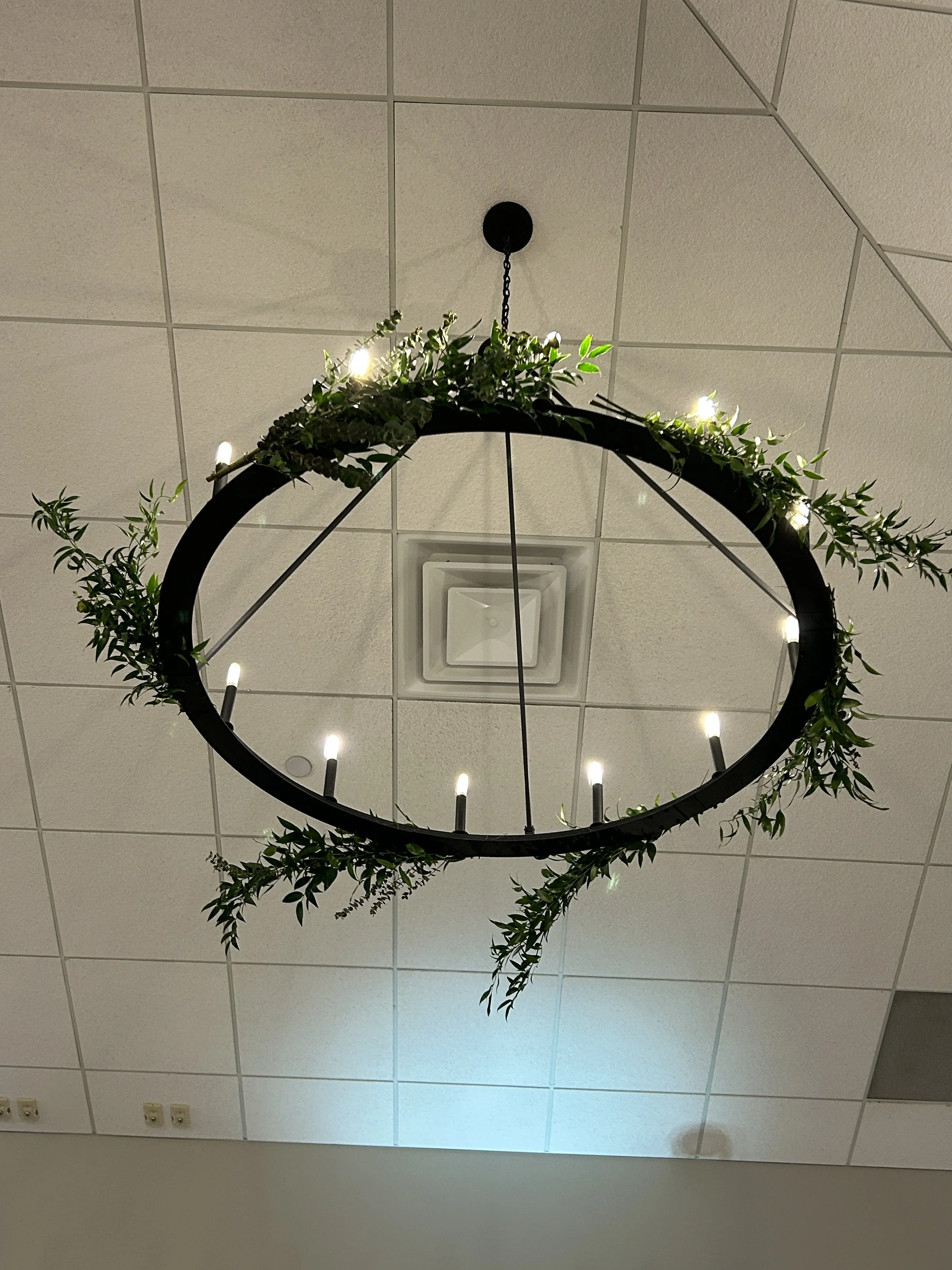 A ceiling with a decorative circular black chandelier wrapped with green foliage and small white lights attached, with a central air vent visible and a grid of ceiling tiles.