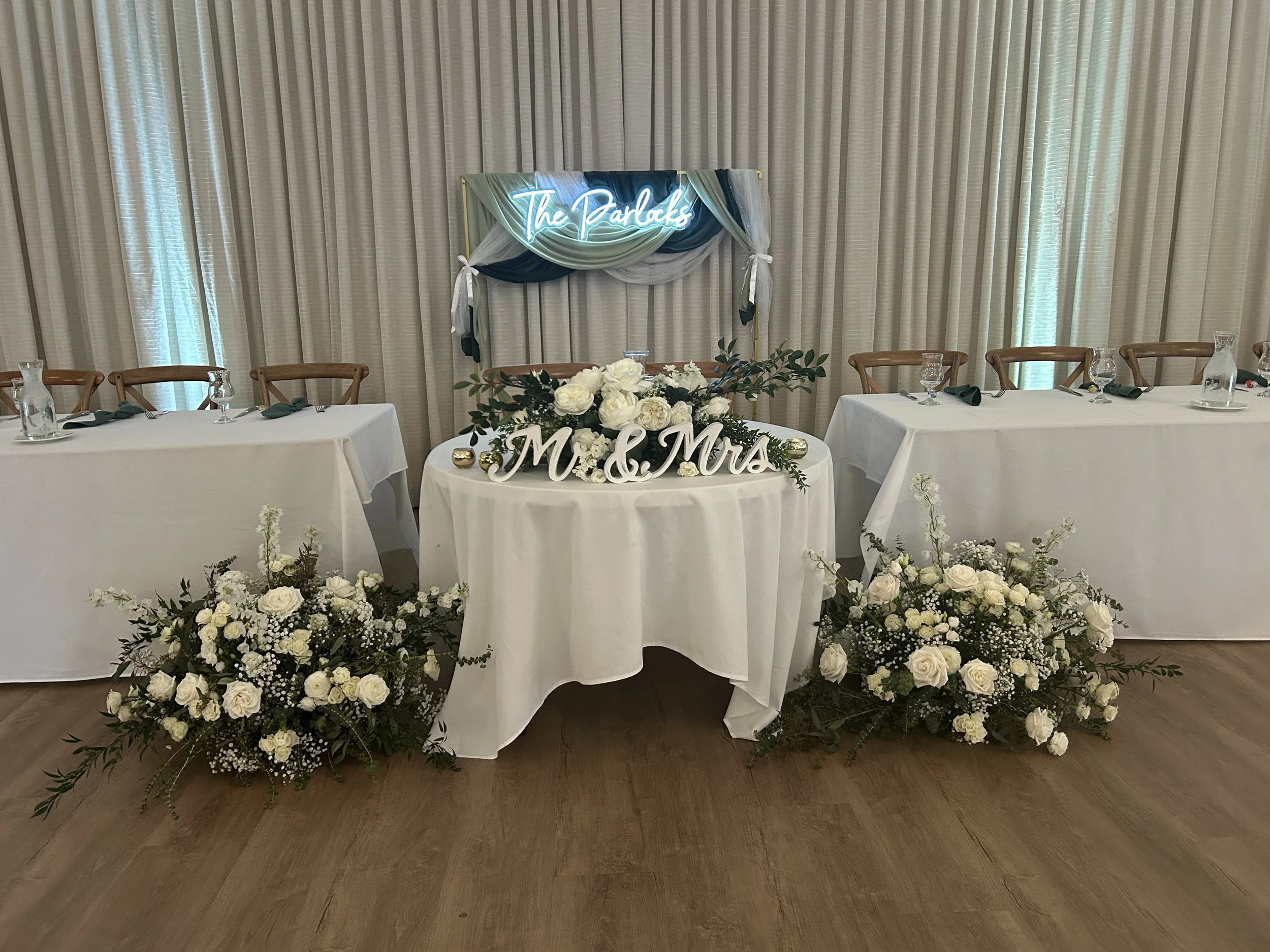 Wedding reception table with floral arrangements, a 'Mr. & Mrs.' sign, and a backdrop with a neon sign reading 'The Parkles'.
