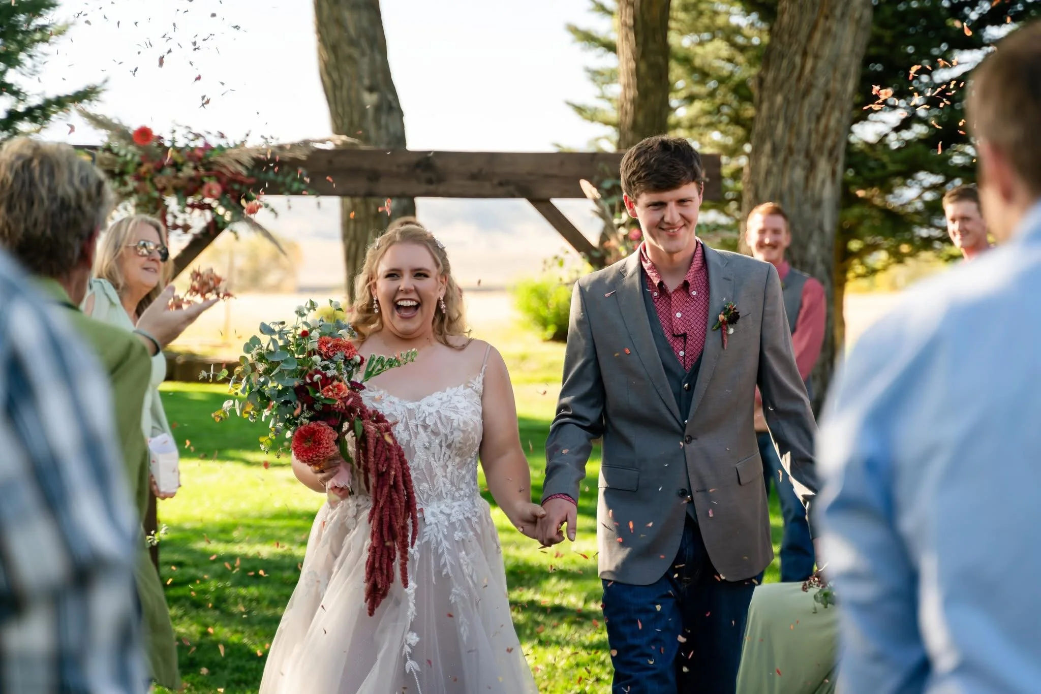 A wedding celebration outdoors with a bride and groom walking hand in hand. The bride is smiling and holding a bouquet, and the groom is smiling. Guests are throwing flower petals around them. There are trees and a wooden arch in the background, and 