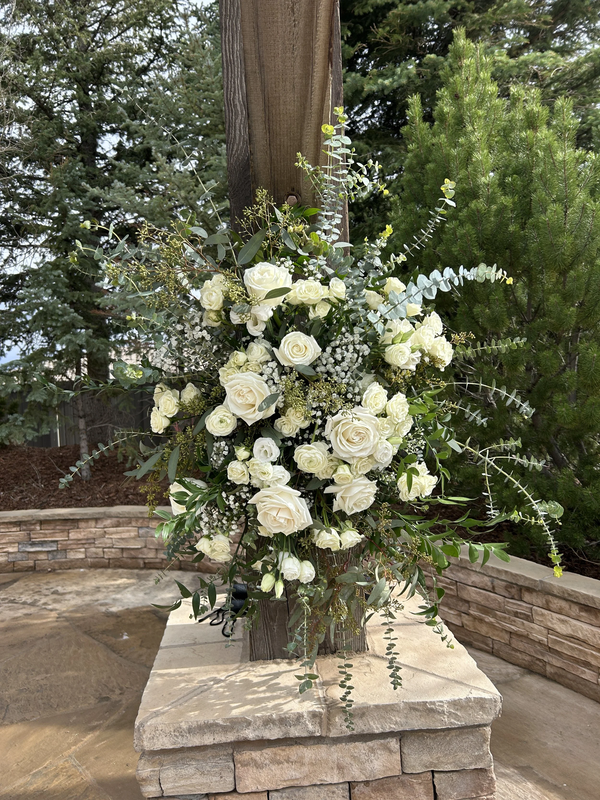 Floral arrangement with white roses, baby's breath, eucalyptus, and other greenery attached to a wooden post outdoors, with trees in the background and a stone garden bed beneath.