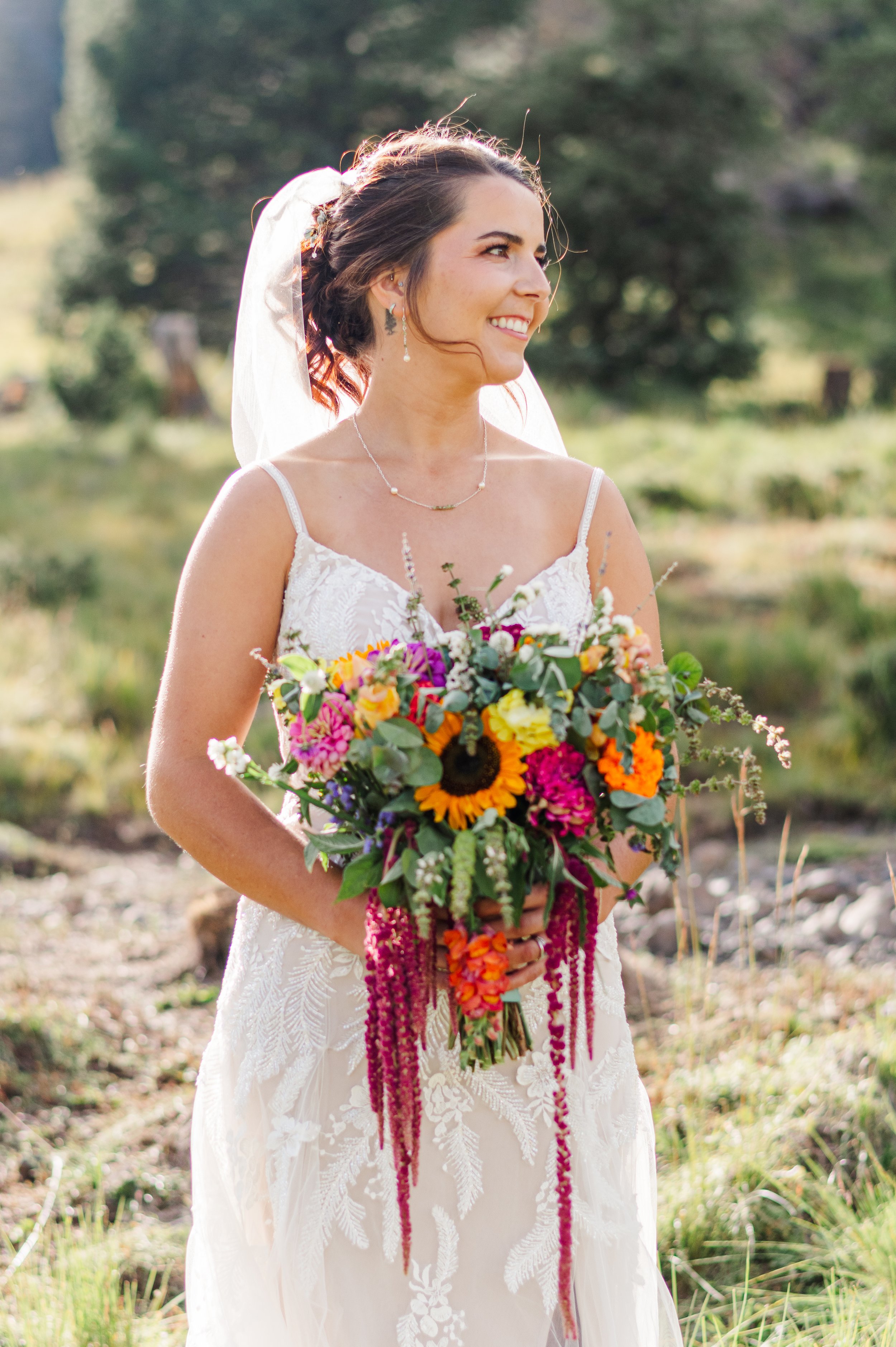 A smiling woman in a wedding dress holding a colorful bouquet outdoors in a field with trees in the background.