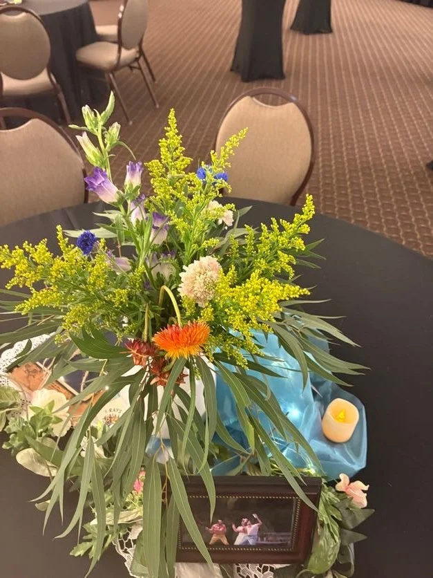 A floral centerpiece with various colorful flowers and green leaves on a black table, with some decor items and chairs in the background.