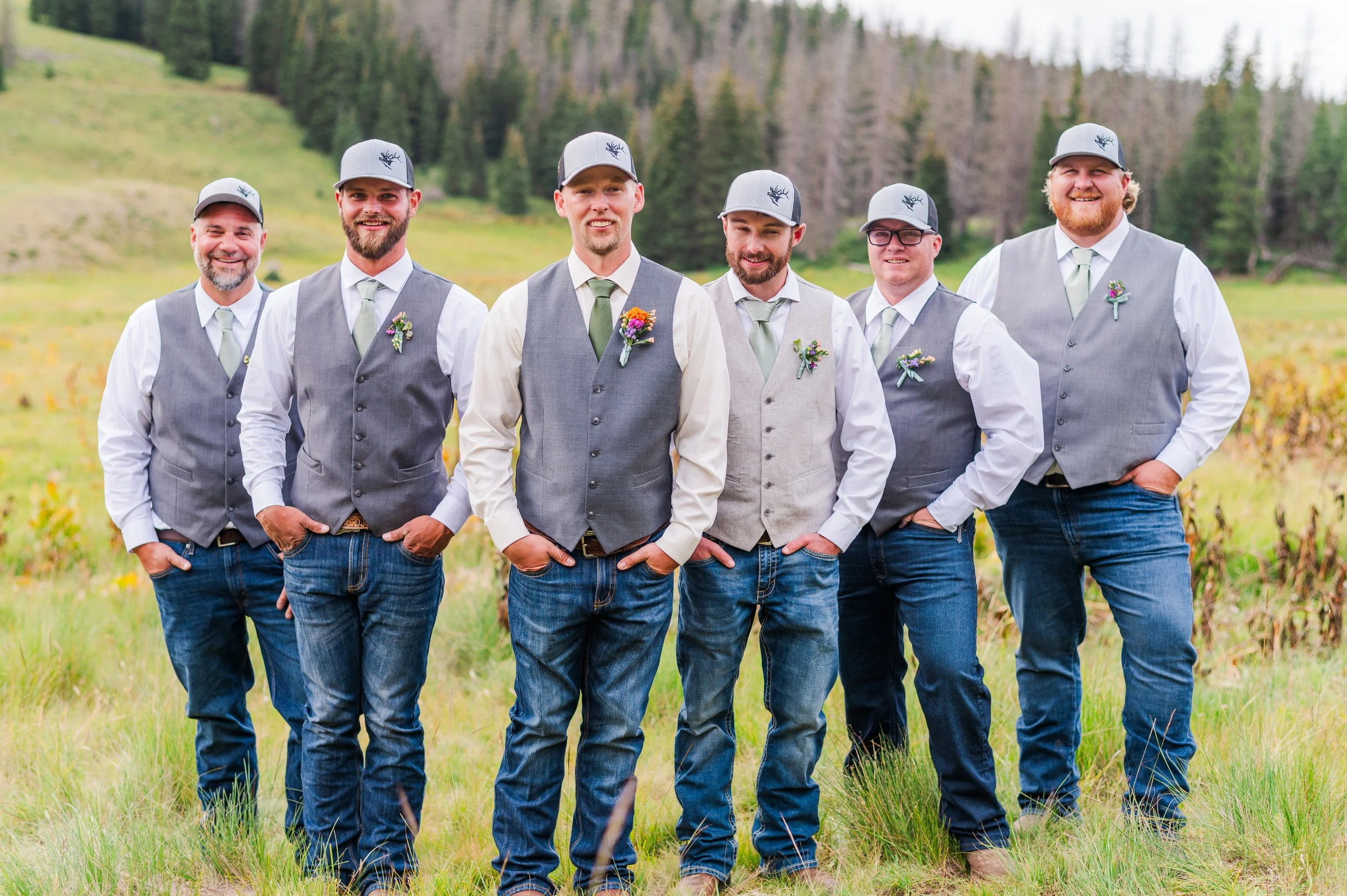 Six men standing outdoors in a field with a forested hillside in the background, dressed in coordinating gray vests, white shirts, green ties, and matching gray caps with a black deer logo. They are smiling and posing for a group photo, with some flo