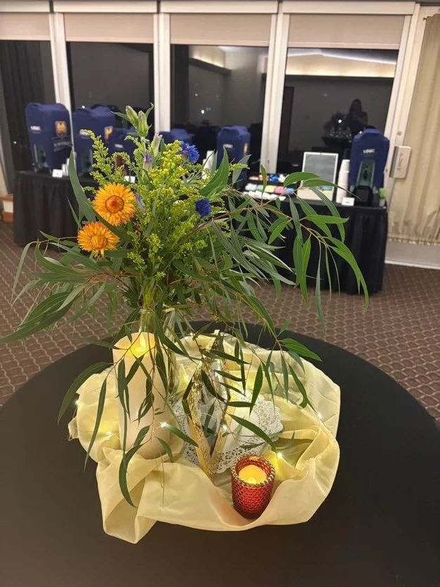 Floral arrangement with yellow, purple, and green flowers and leaves on a table with candles and decorations, in a room with a window and a DJ setup in the background.