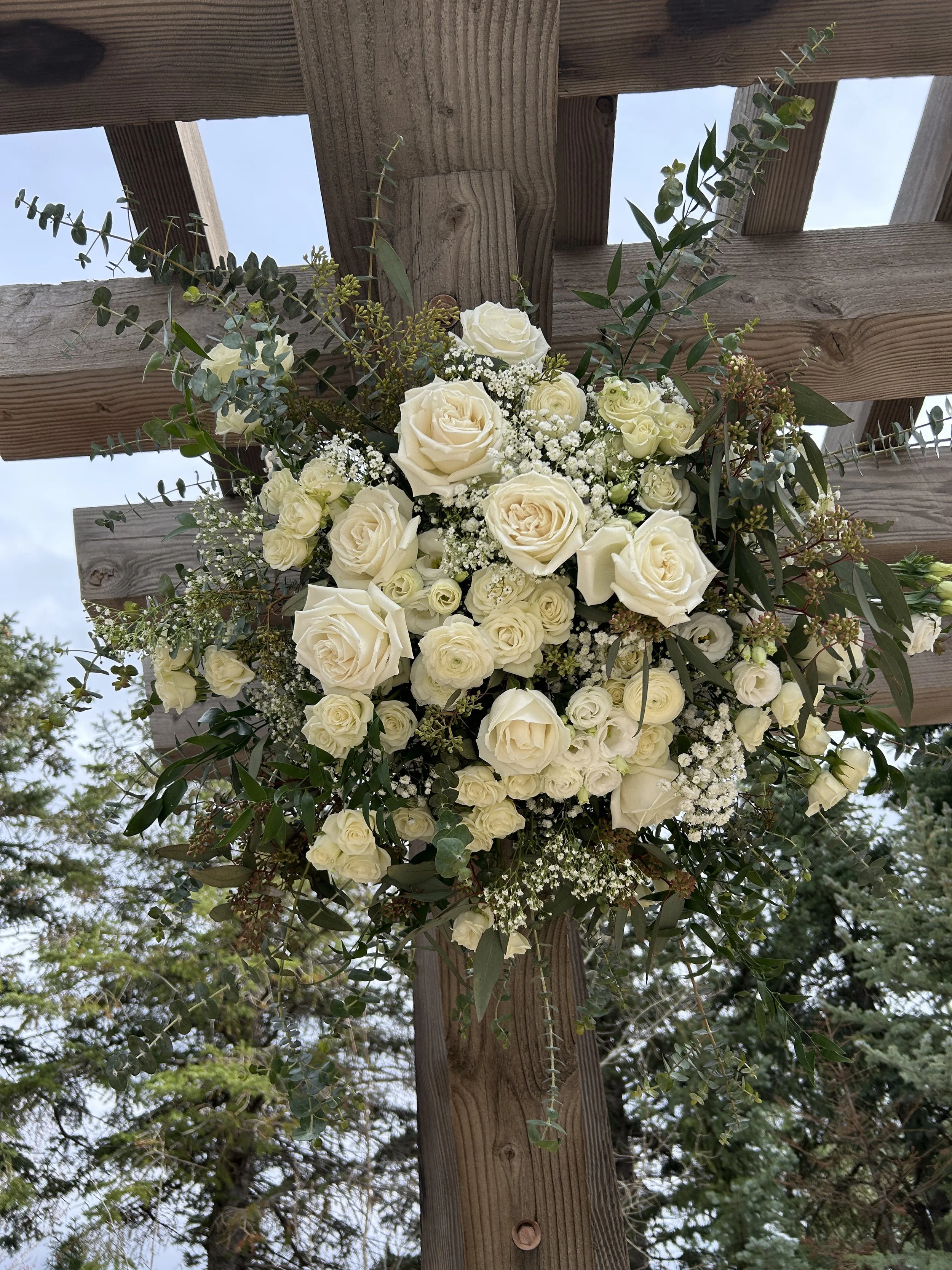 A bouquet of white roses and baby's breath with green foliage, attached to a wooden structure with screws, against a background of trees and blue sky.