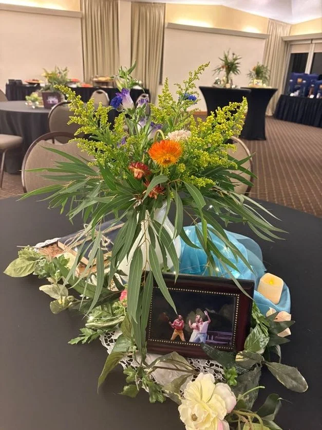 A flower arrangement with green leaves, orange and purple flowers on a round table decorated with a photo frame, candles, and decorative greenery, in a banquet hall setting.