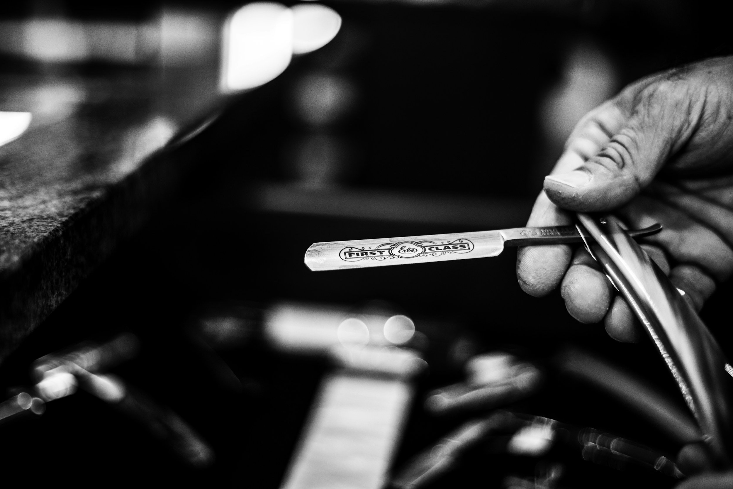 Close-up of a man's hand holding a straight razor with the label 'First Class' in a barbershop.