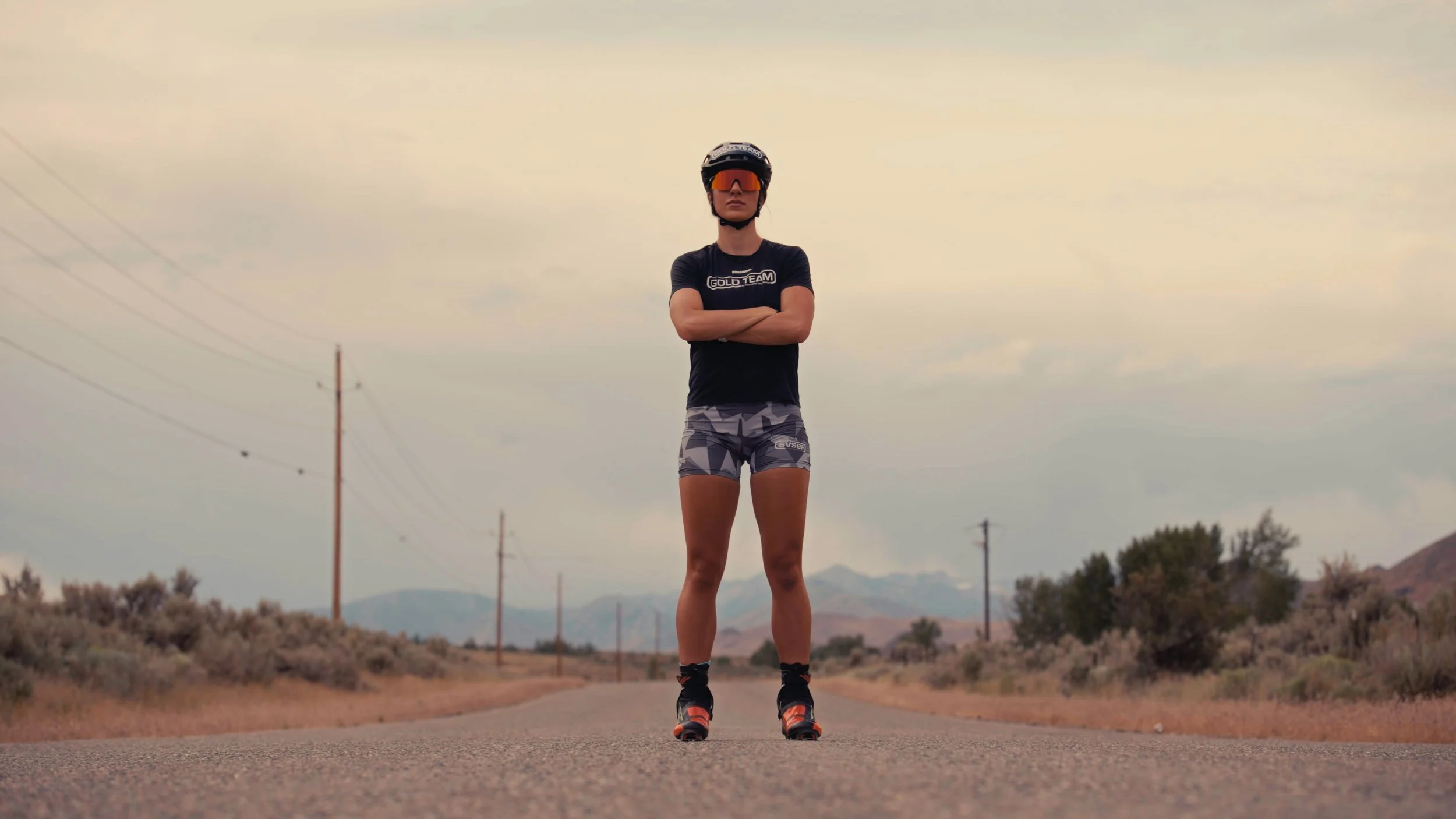 A woman standing on an empty road in the outdoors, wearing a black helmet, orange sunglasses, black T-shirt, camouflage shorts, and running shoes, with arms crossed and a mountain landscape in the background.