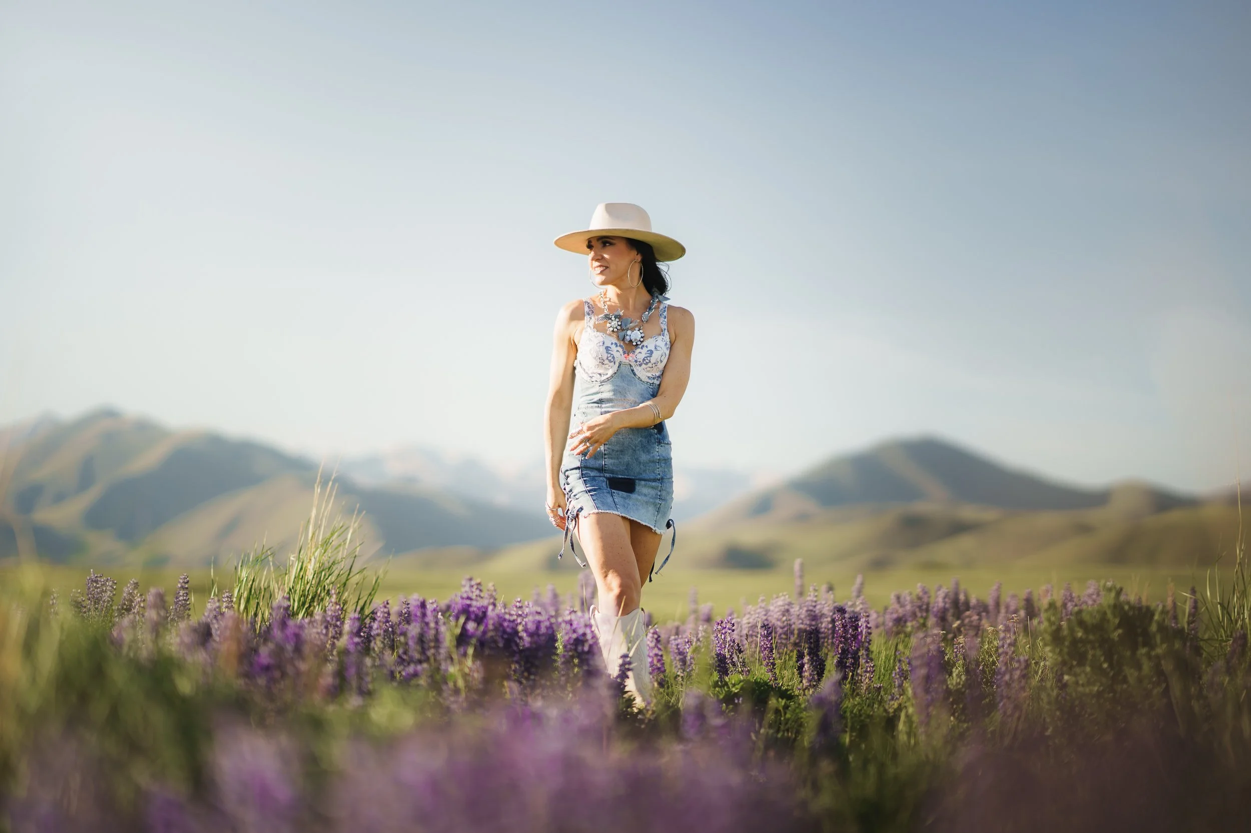 A woman wearing a wide-brimmed hat, a floral top, and denim shorts standing in a field of purple flowers with mountains in the background.