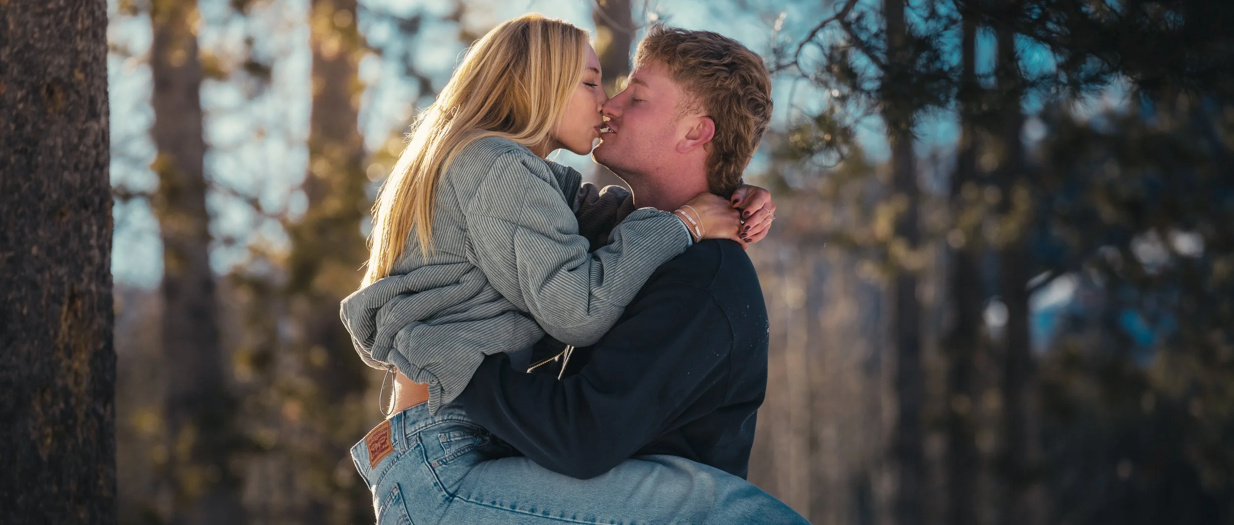 A young couple kissing outdoors in a wooded area during daytime, with the girl sitting on the boy's lap and the boy holding her around the waist.