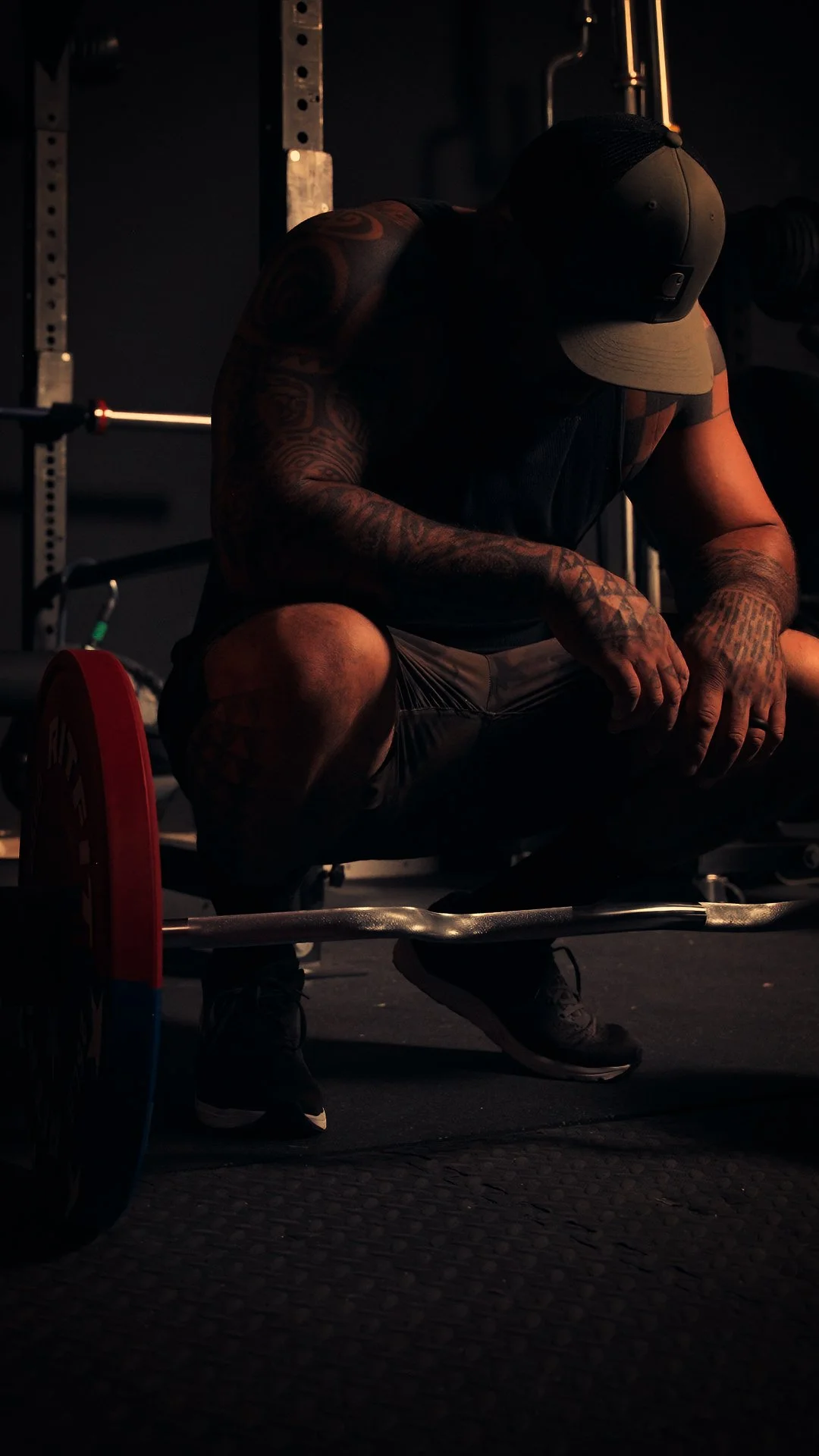 A muscular man sitting in a dark gym, wearing a black cap and black sports attire, with tattoos on his arms, possibly resting after lifting weights.