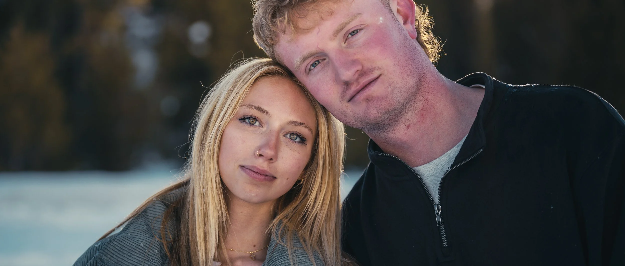 A young man and woman with light skin and blond hair outdoors, close together, with a blurred natural background.
