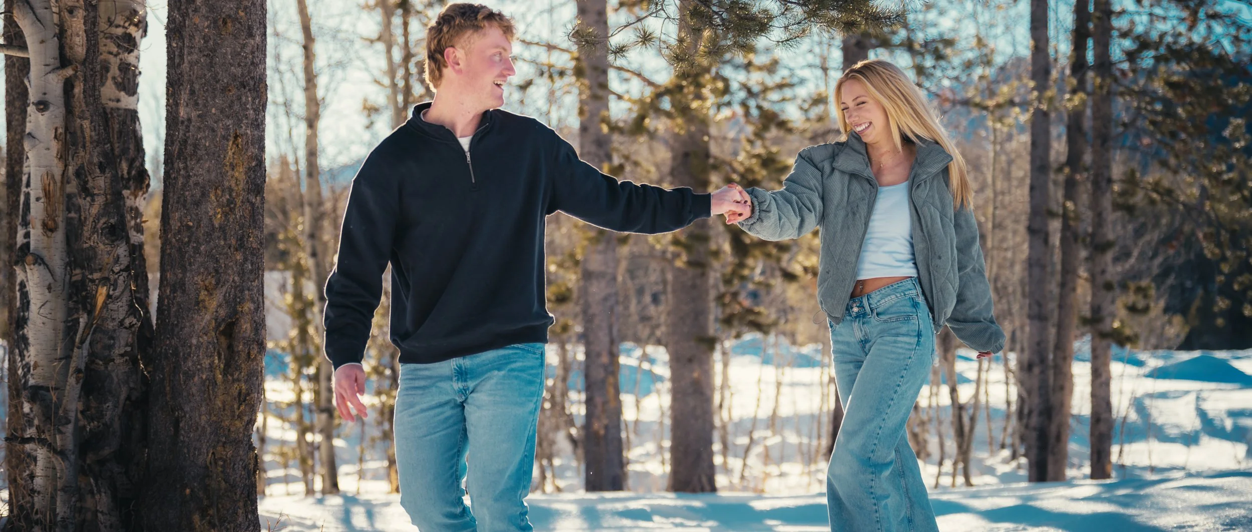 A young man and woman smiling and holding hands in a snowy forest, enjoying a walk in winter.