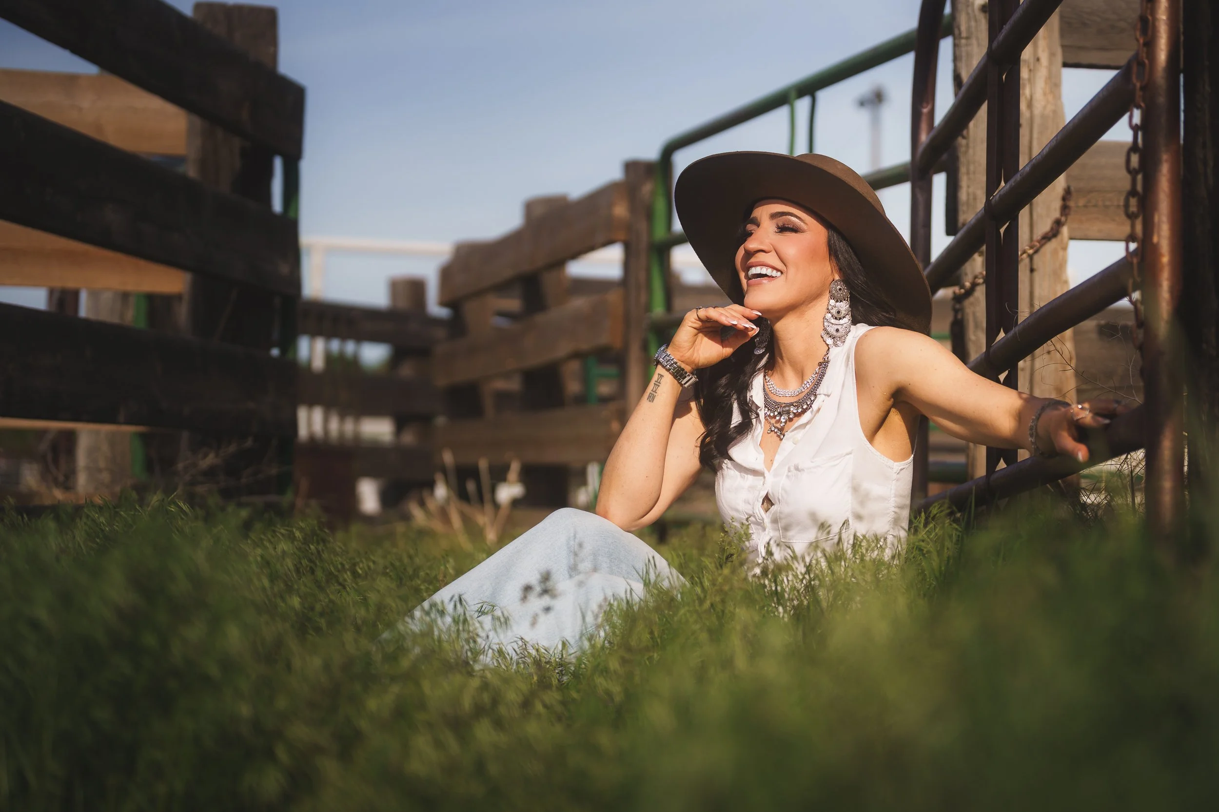 A woman sitting on the grass near a wooden and metal structure outdoors, smiling and wearing a wide-brimmed hat, large earrings, and layered necklaces.