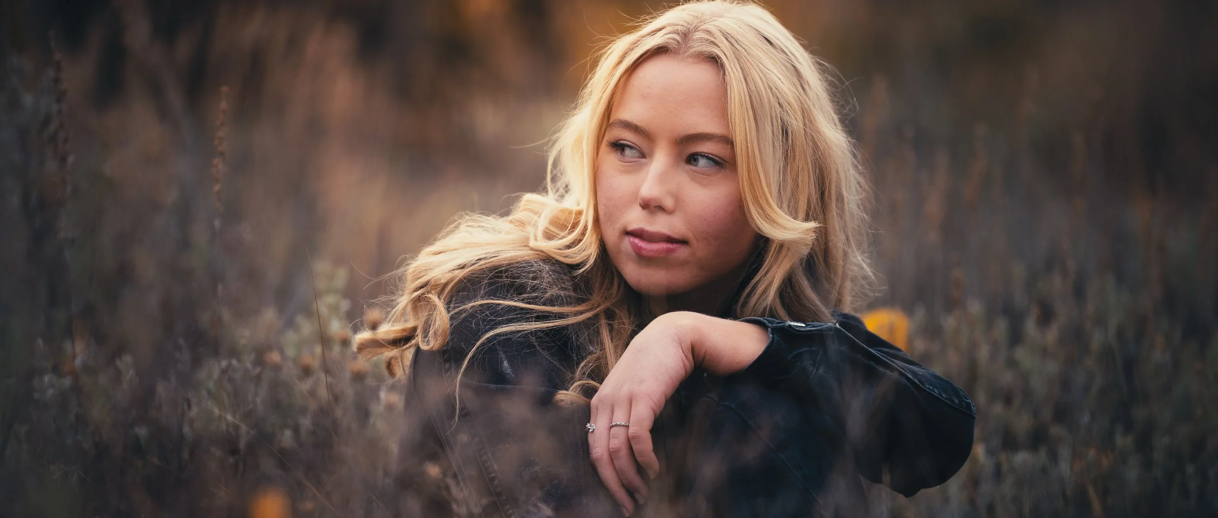 A young woman with long blonde hair, wearing a black jacket, sitting outdoors in a grassy area during sunset, looking to her left with a thoughtful expression.