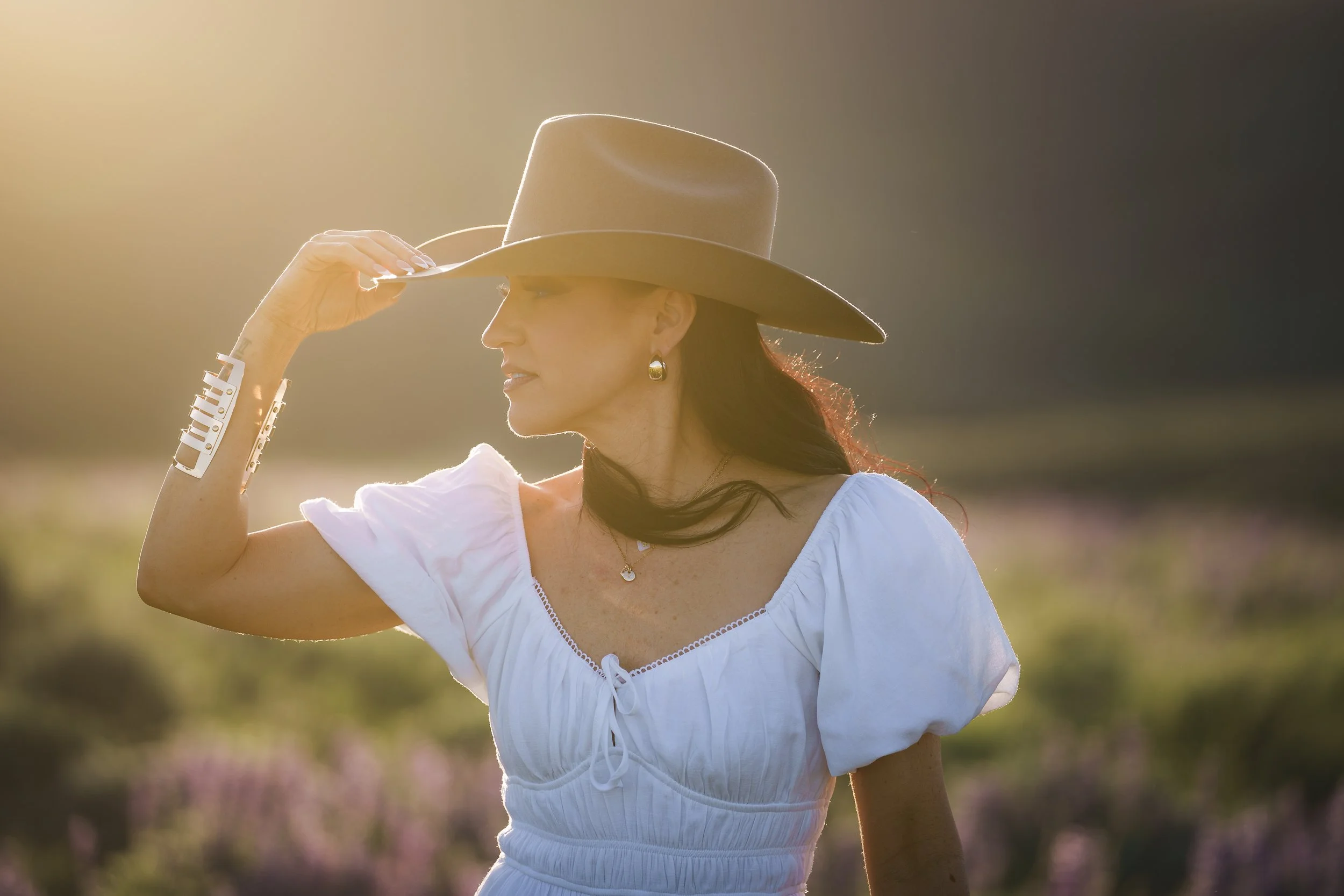 A woman with dark hair wearing a beige wide-brimmed hat, white dress with ruffled sleeves, and jewelry, standing outdoors in a sunny field, adjusting her hat with one hand.