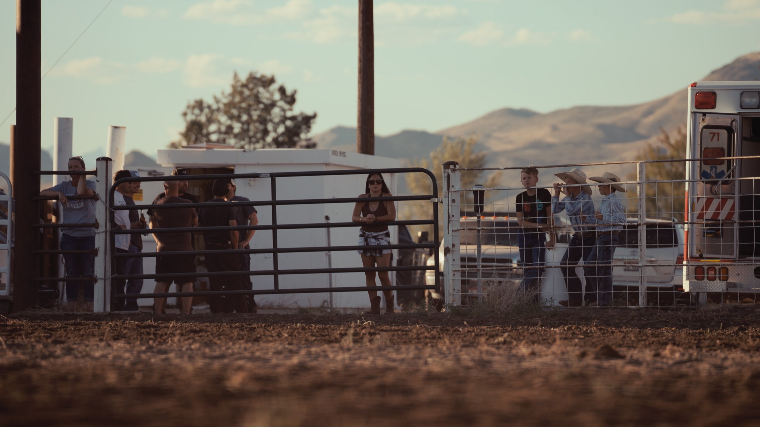 A group of people standing behind a metal fence at an outdoor event, with mountains and a clear sky in the background.