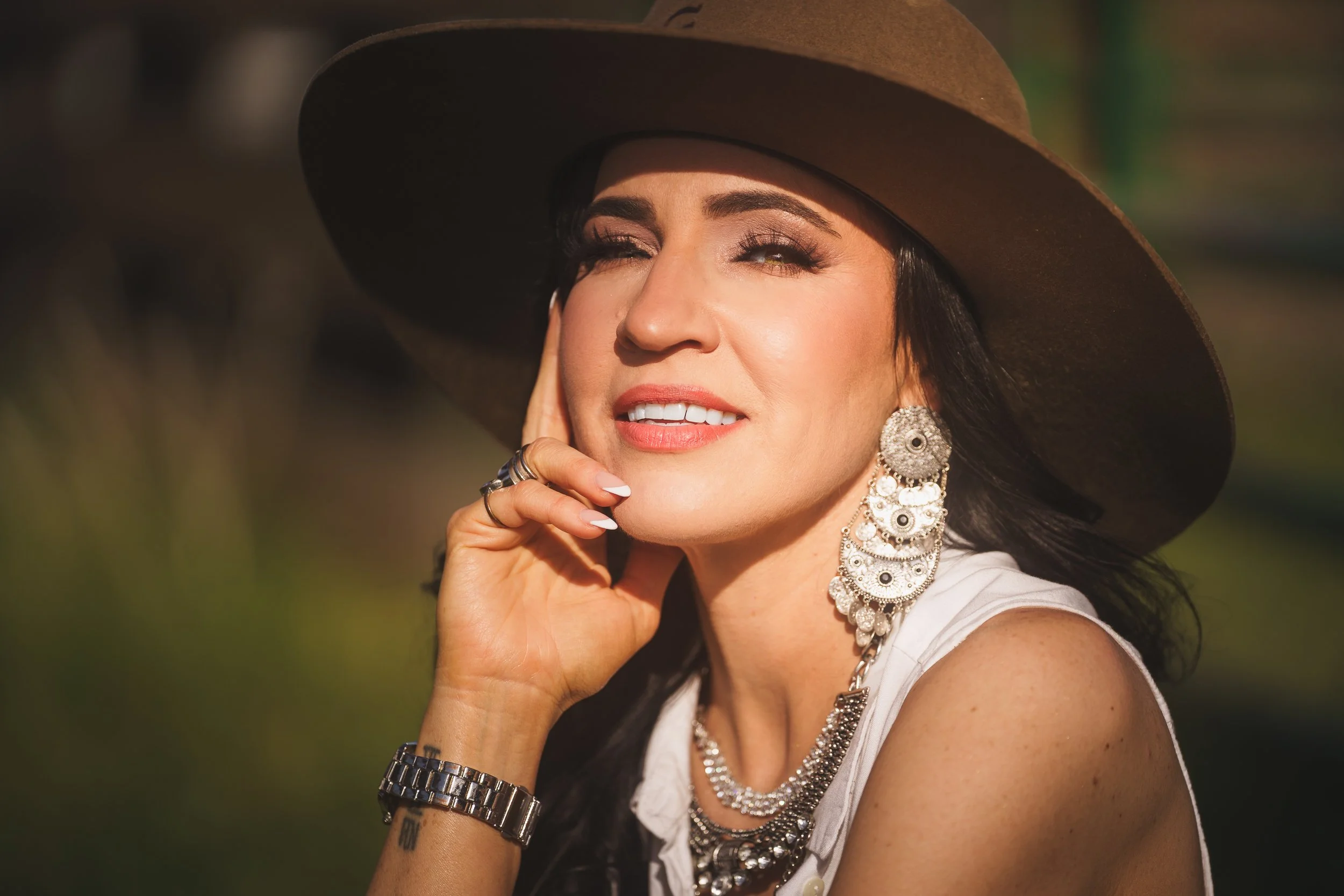A woman wearing a large wide-brimmed brown hat and ornate jewelry, including large earrings, necklaces, and rings, smiling and resting her chin on her hand outdoors.