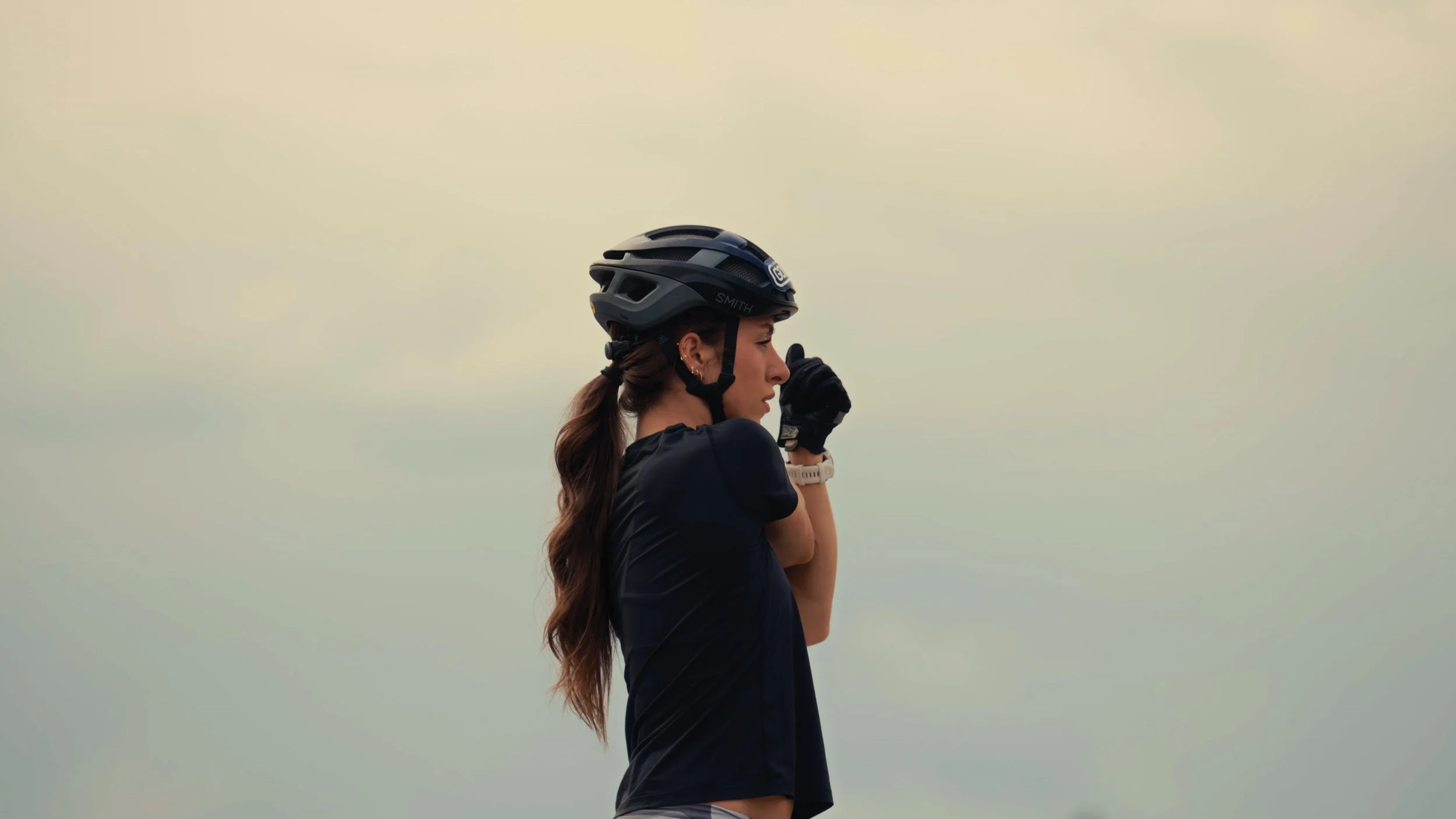 A woman wearing a black cycling helmet and gloves adjusting her helmet against a pale sky.