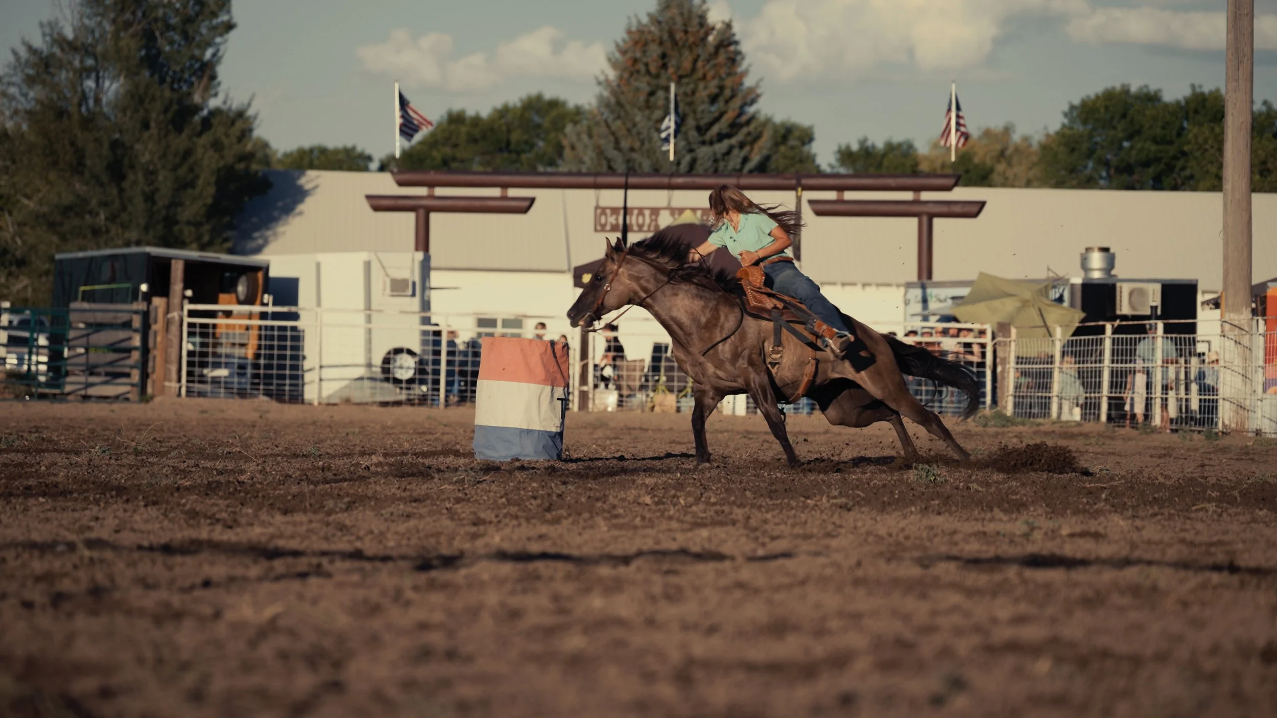 A woman riding a horse in a dirt arena, leaning into a turn around a barrel during a rodeo or horse riding event, with fences, trailers, flags, and spectators in the background.