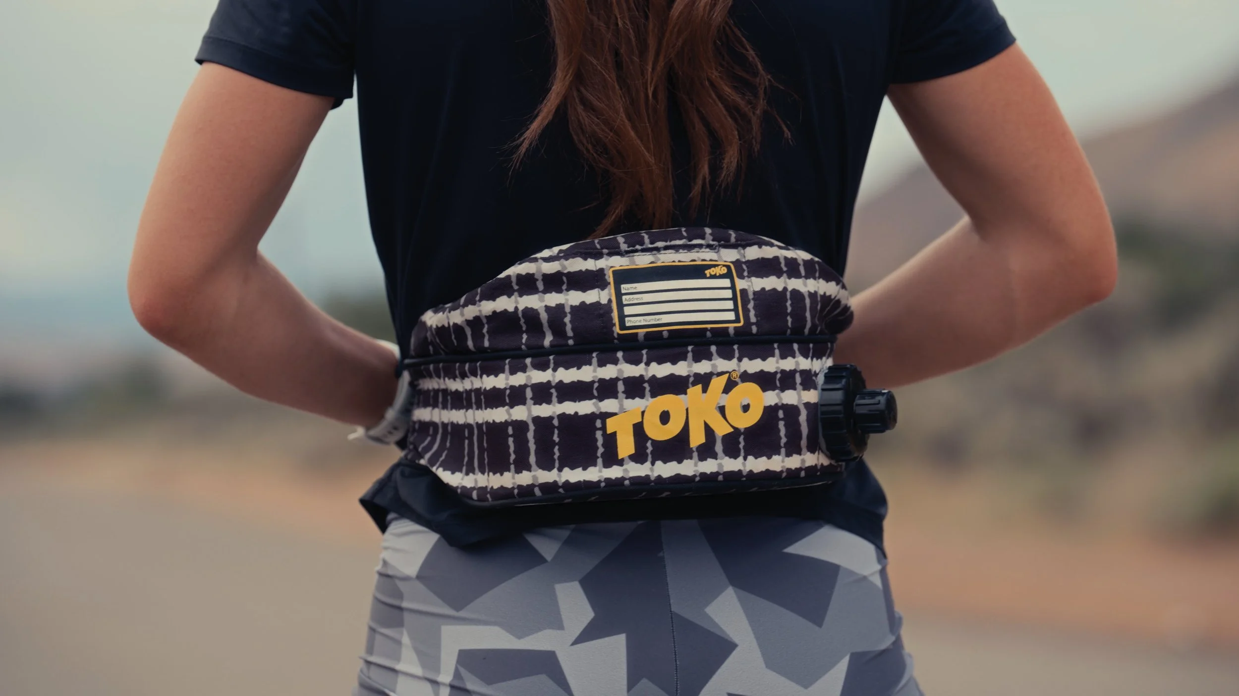 A person wearing a black shirt and patterned gray shorts, carrying a black and white patterned waist pack with the word 'TOKO' in yellow, standing outdoors.