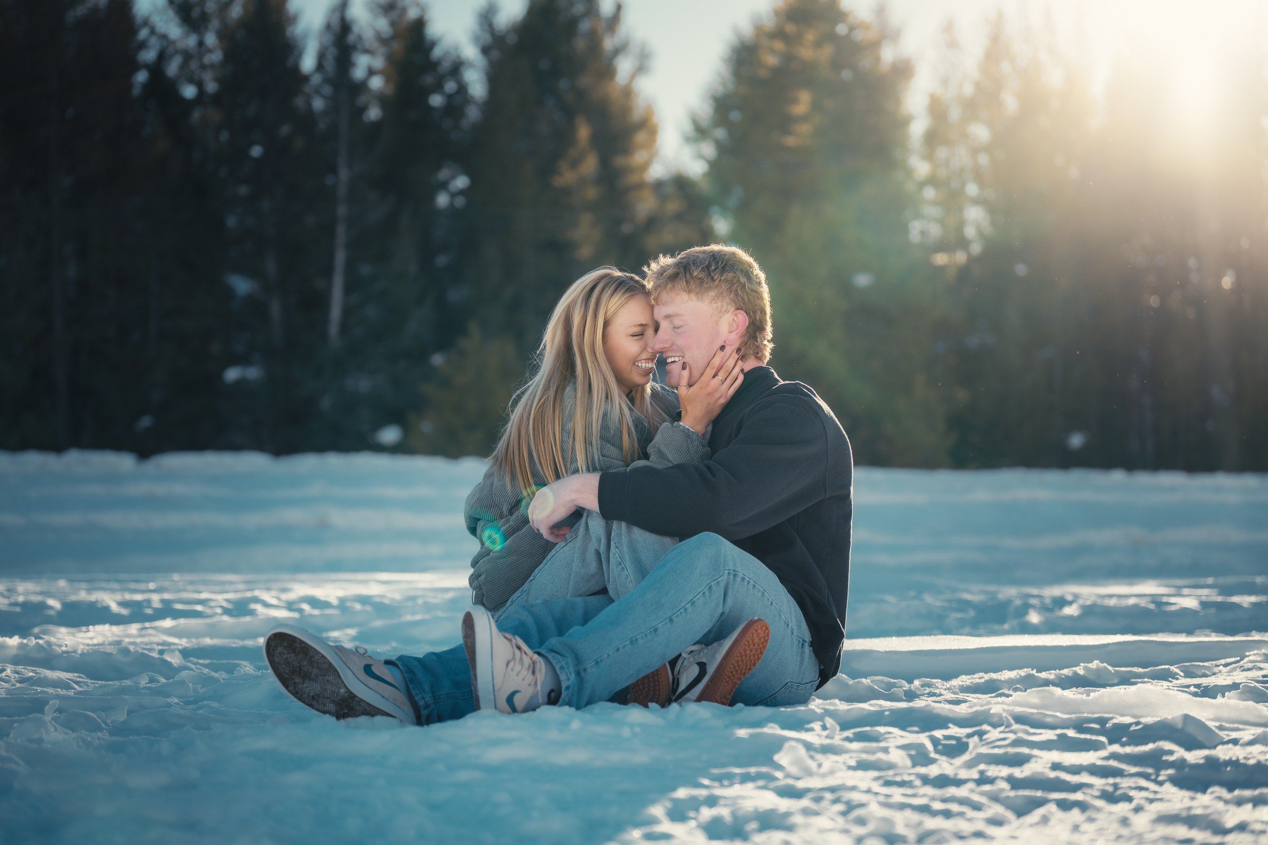 A young couple sitting on snow-covered ground in a forest, smiling and leaning close to each other, with sunlight filtering through trees in the background.