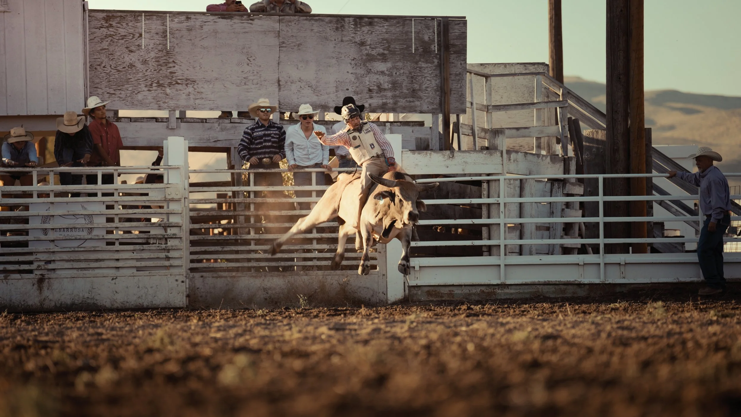 A rodeo scene with a cowboy riding a bucking bull within a fenced arena, spectators watching from a wooden platform.