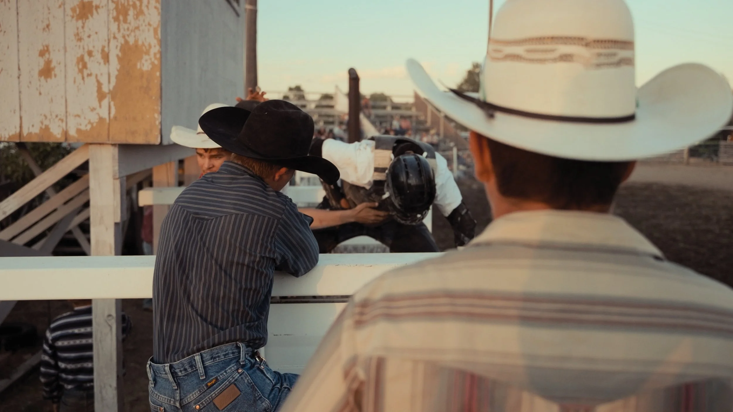 Group of people wearing cowboy hats gathered outdoors, with some leaning on a white railing, and a small building in the background under a clear sky during sunset.