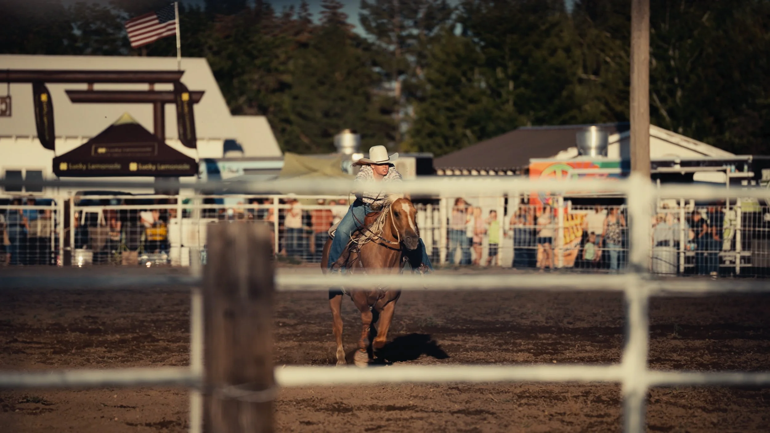 A rodeo event with a woman riding a horse inside a fenced arena, wearing a white cowboy hat and patterned shirt, with spectators and structures in the background.