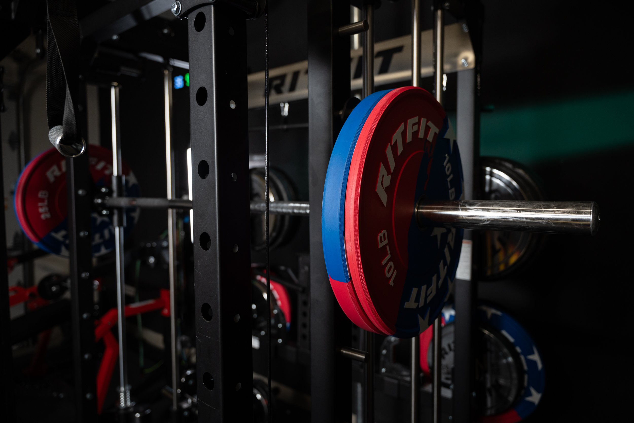 Close-up of a power rack with red and blue weight plates labeled 'RIT FIT' on a barbell in a gym.