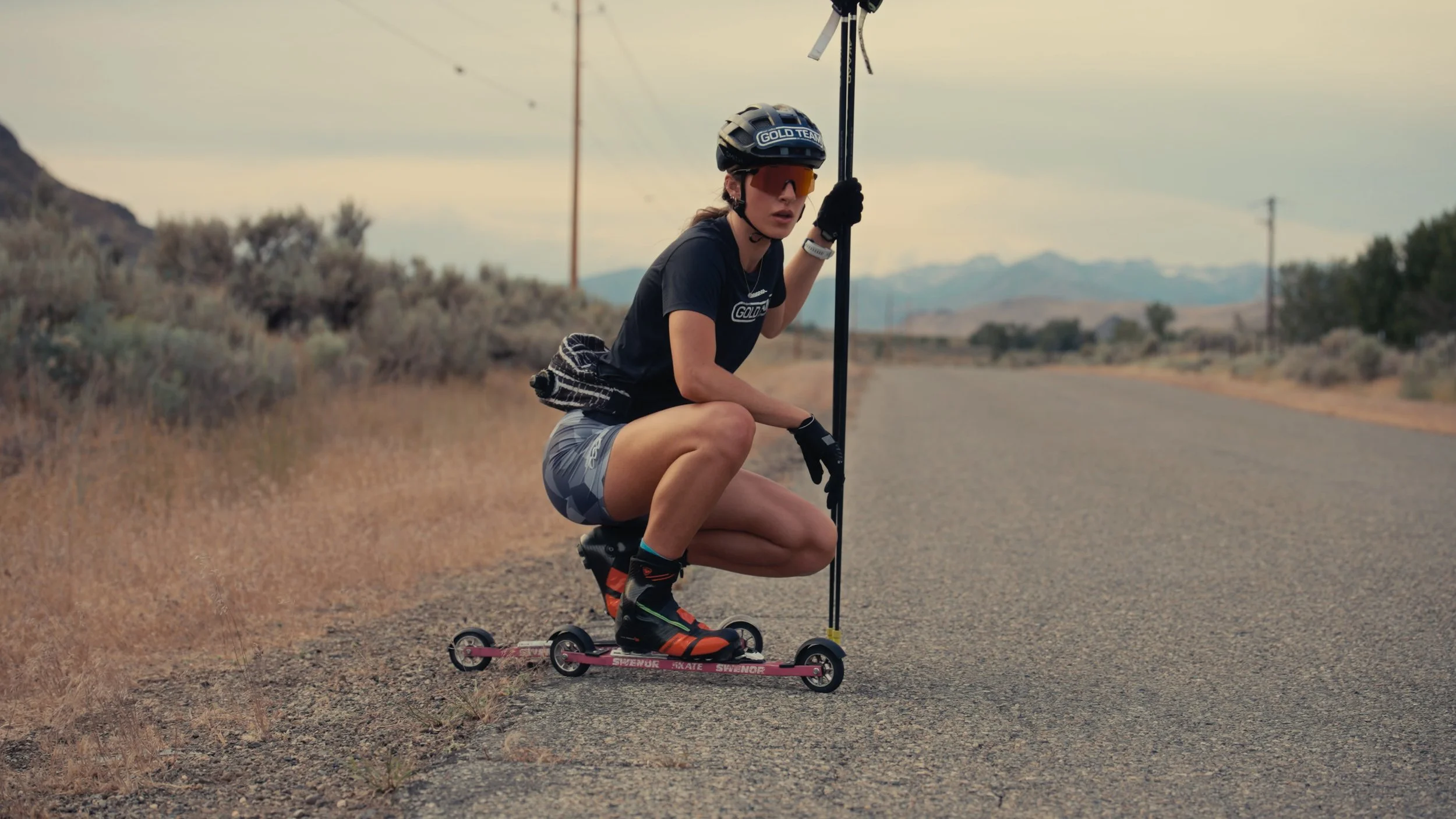 A young woman wearing a helmet and sunglasses crouching on a skateboard on a desert road, holding a ski pole, with mountains and power lines in the background.