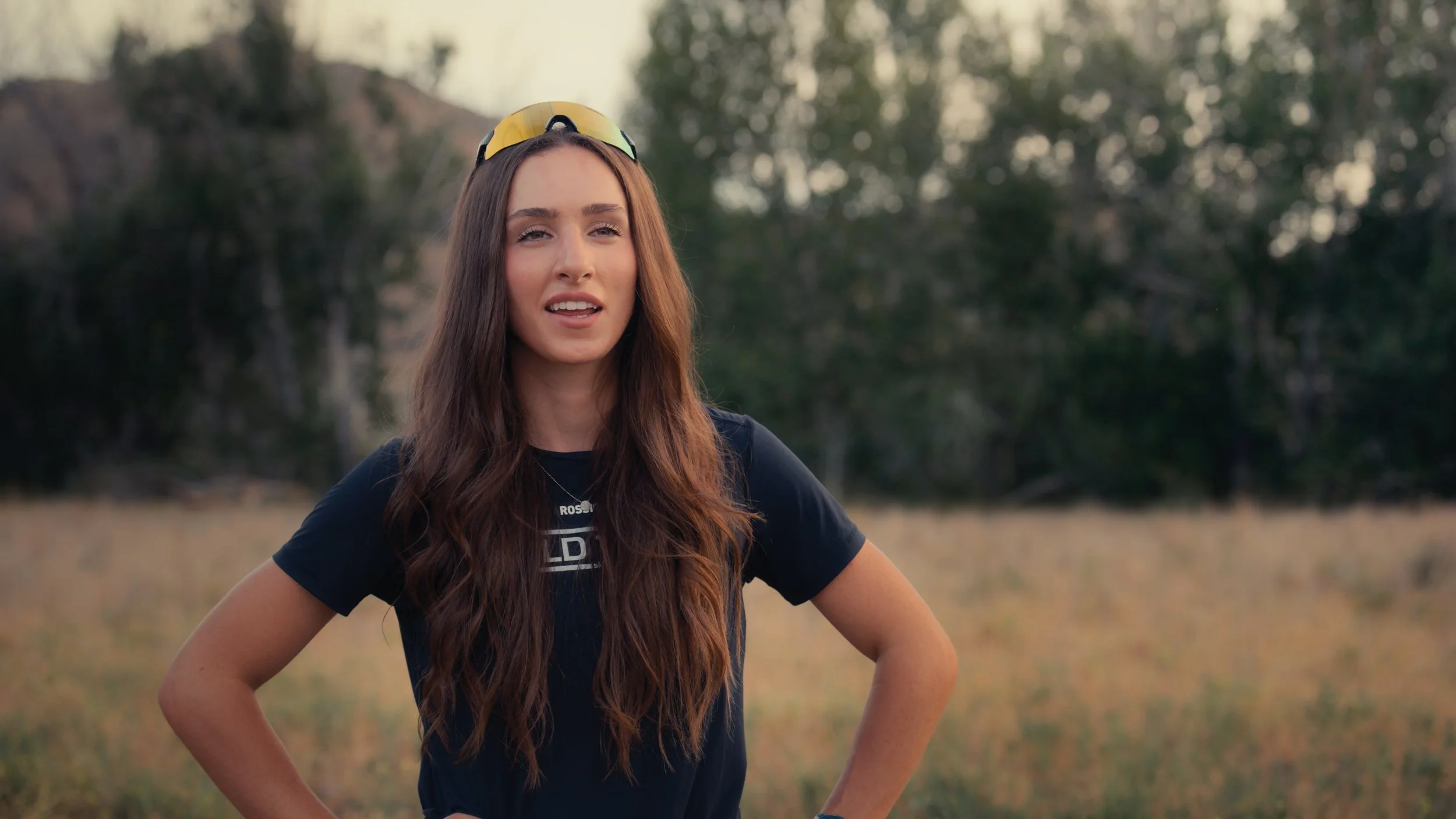 A young woman with long brown hair wearing a black t-shirt and yellow sunglasses on her head standing outdoors with a blurred background of trees and mountains.