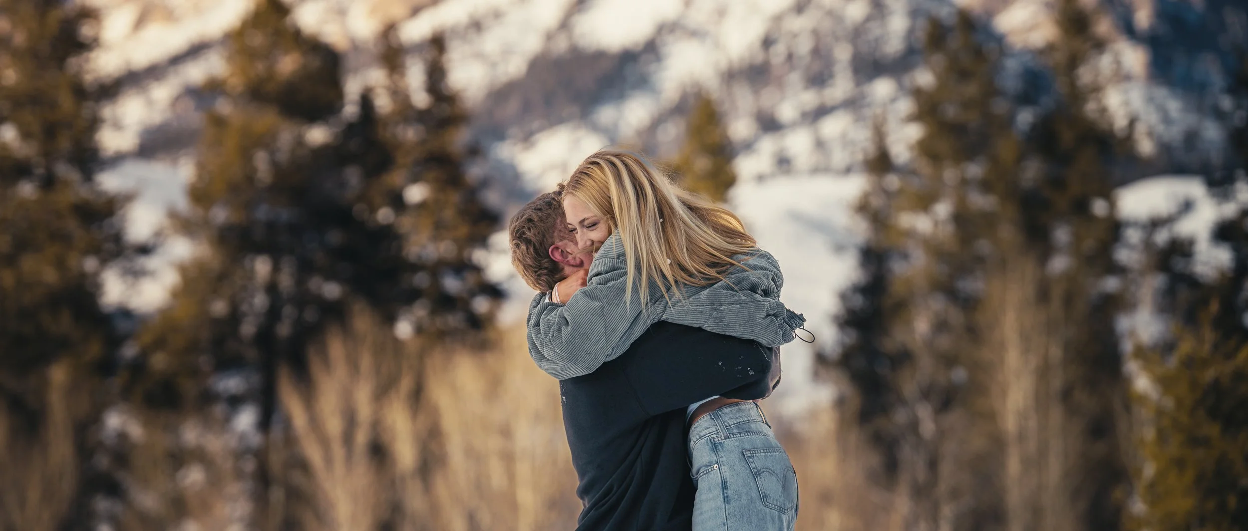 A couple embracing outdoors in a snowy mountainous area, the woman smiling with arms around the man's neck, the man lifting her with snow-covered trees and mountains in the background.