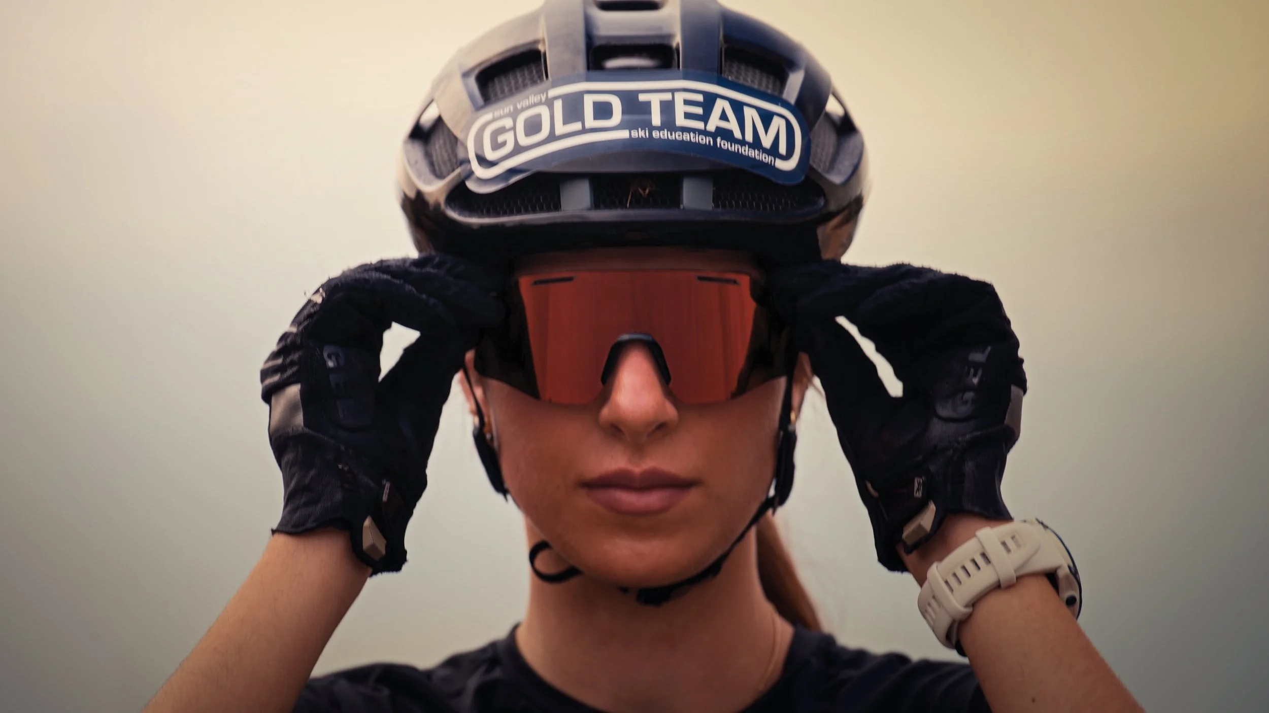 Roller Ski Person wearing a black helmet with a label that reads 'GOLD TEAM,' adjusting orange goggles while wearing black gloves.