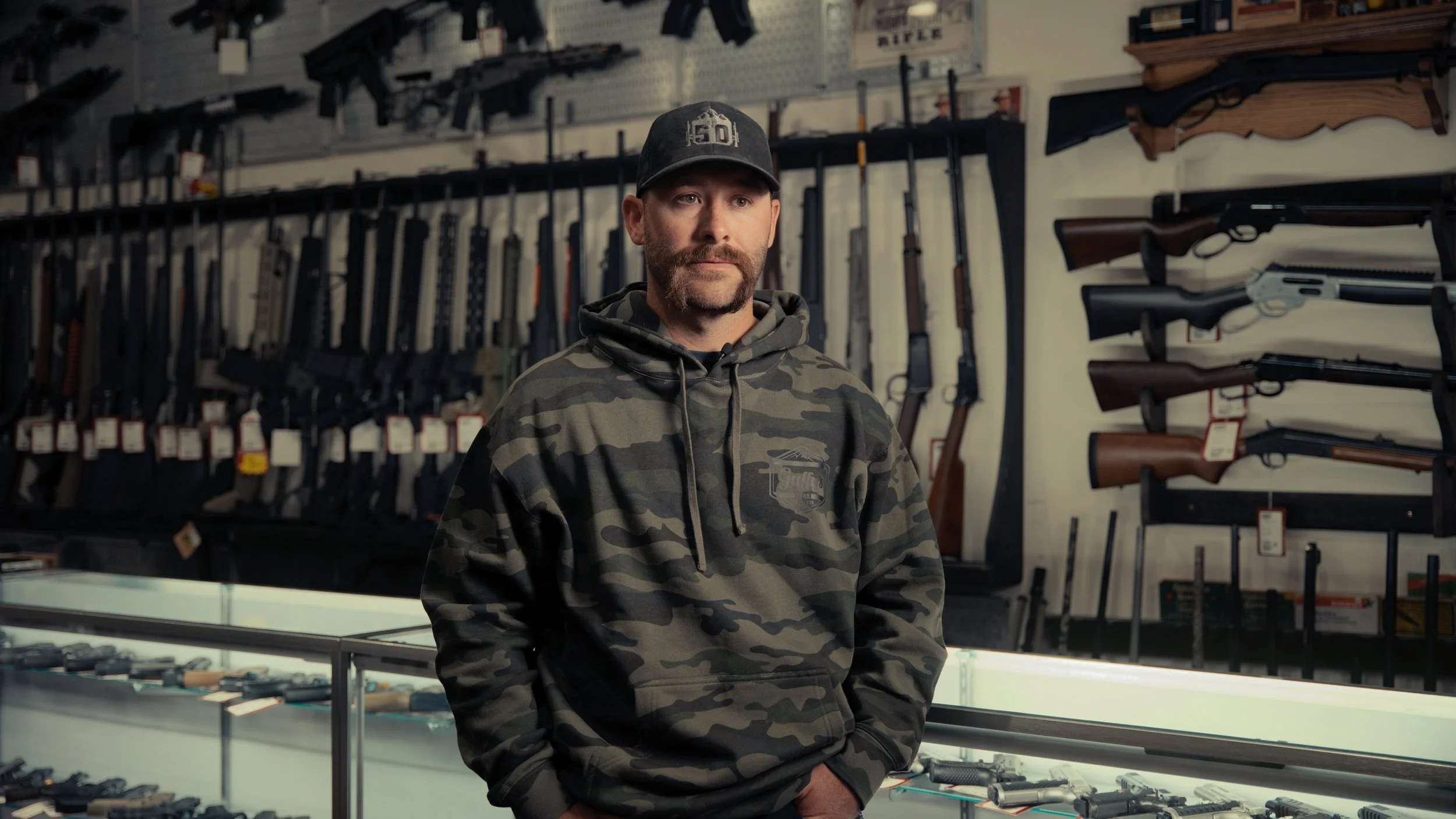 A man wearing a camouflage hoodie and a black baseball cap stands inside a gun store, with rifles displayed on the wall behind him.