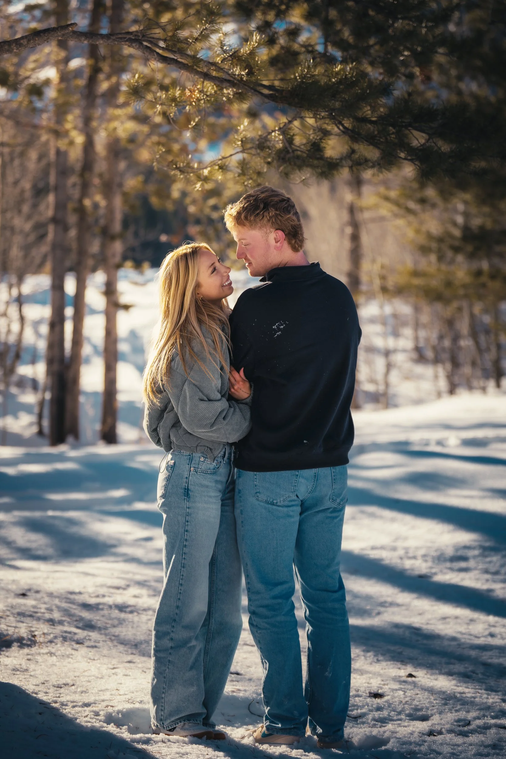 A young couple stands close together in a snowy outdoor setting, with sunlight filtering through the trees. They are smiling and looking into each other's eyes, appearing happy and affectionate.