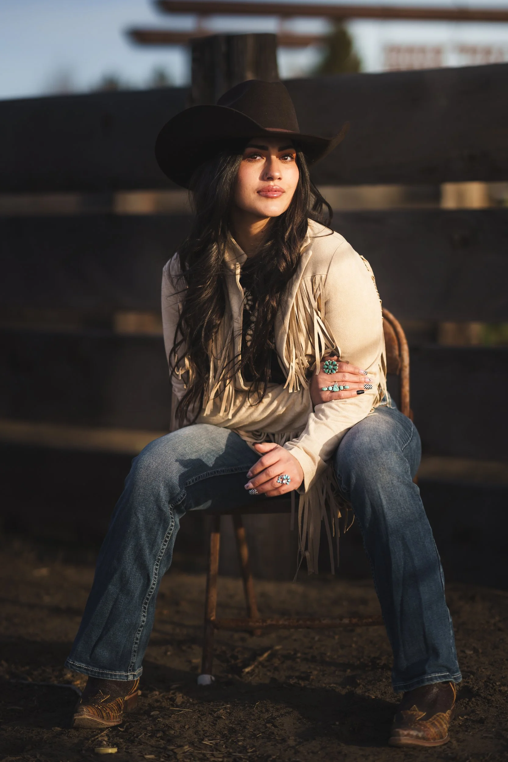 A woman wearing a black cowboy hat, a tan fringed jacket, blue jeans, and cowboy boots, sitting on a wooden chair outdoors during late afternoon.