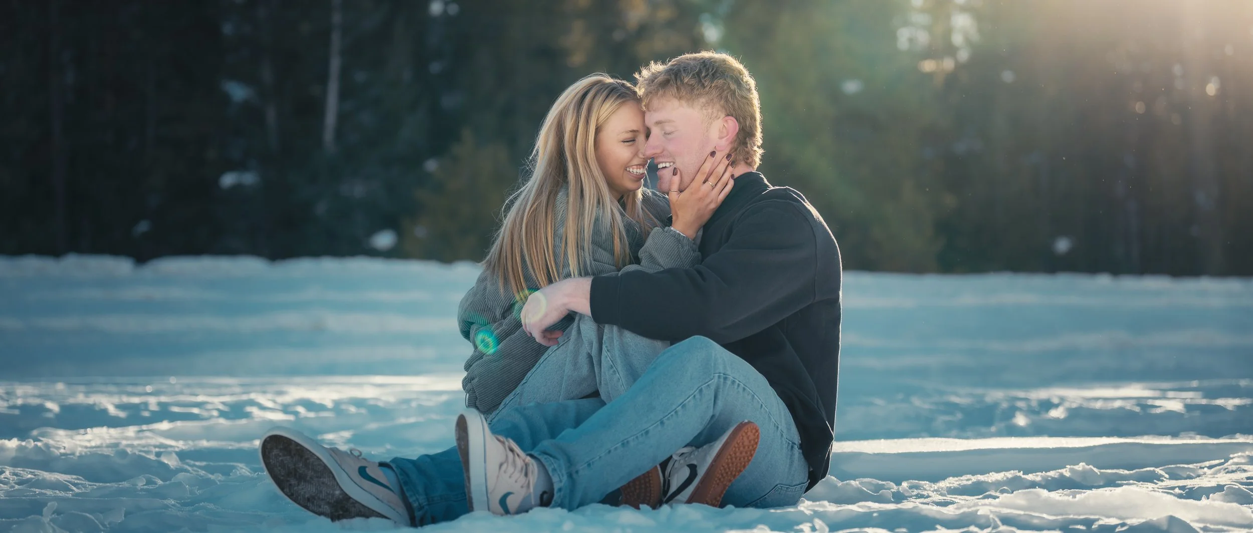 A young man and woman sitting on snow-covered ground in a forest, smiling and about to kiss, with sunlight shining through trees in the background.