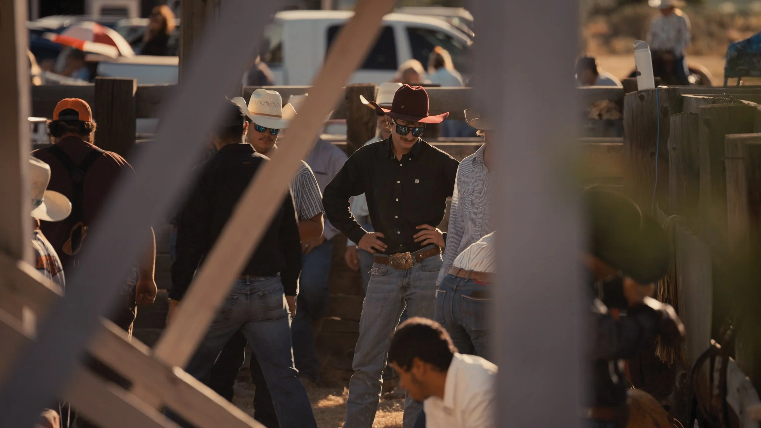 Group of cowboys wearing hats and sunglasses standing and talking at a rodeo or country event, with some people and vehicles visible in the background during sunset.