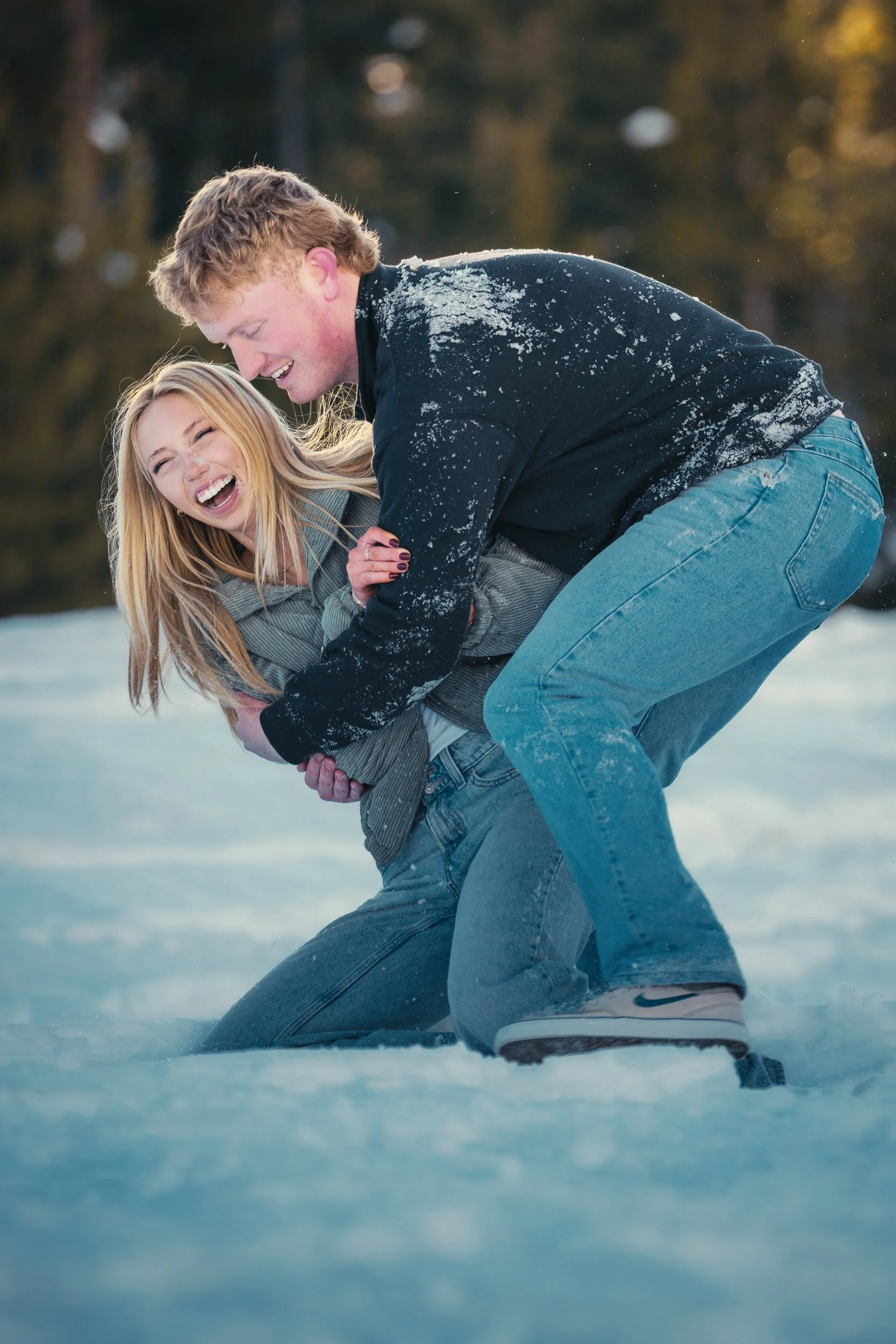 A young couple playing and laughing on the snow outdoors during daytime, with a background of blurred trees and a warm sunset glow.