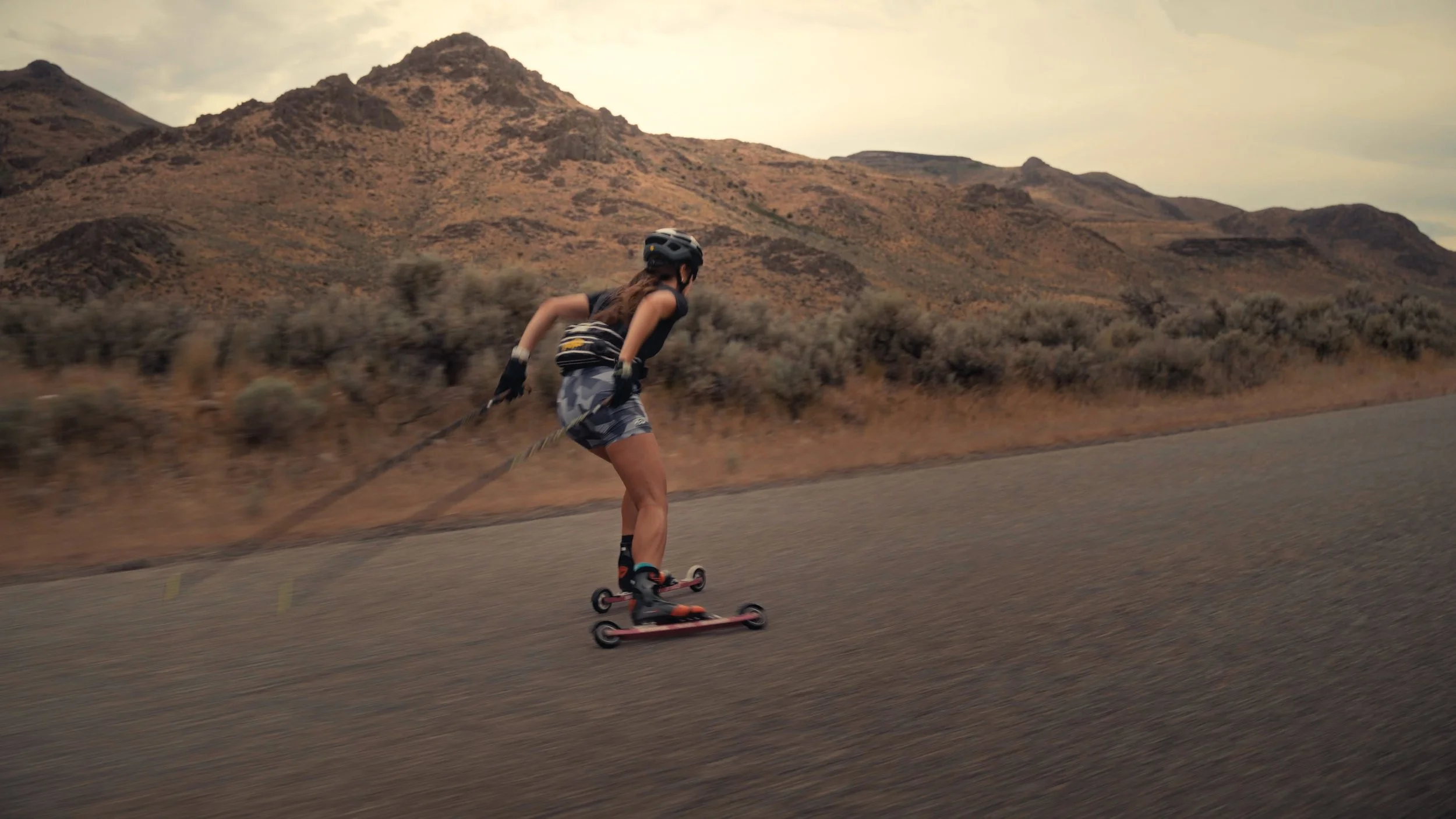 A person wearing a helmet, gloves, and sporty clothing is riding a roller skis downhill on a paved road in a desert landscape with mountains in the background.